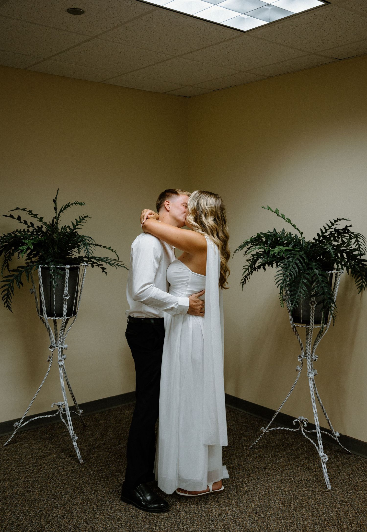 Newlyweds share a romantic first kiss in a cozy indoor ceremony space flanked by lush green ferns.