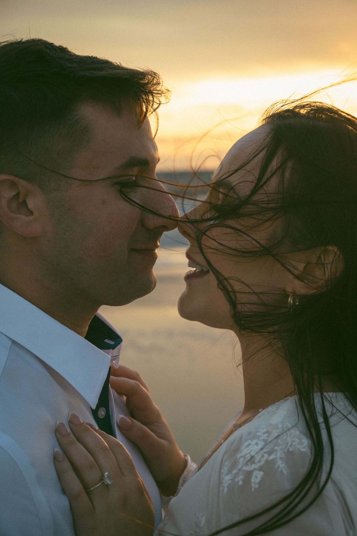 Romantic couple nearly kissing at sunset on a beach, bride's hair blowing in the wind.