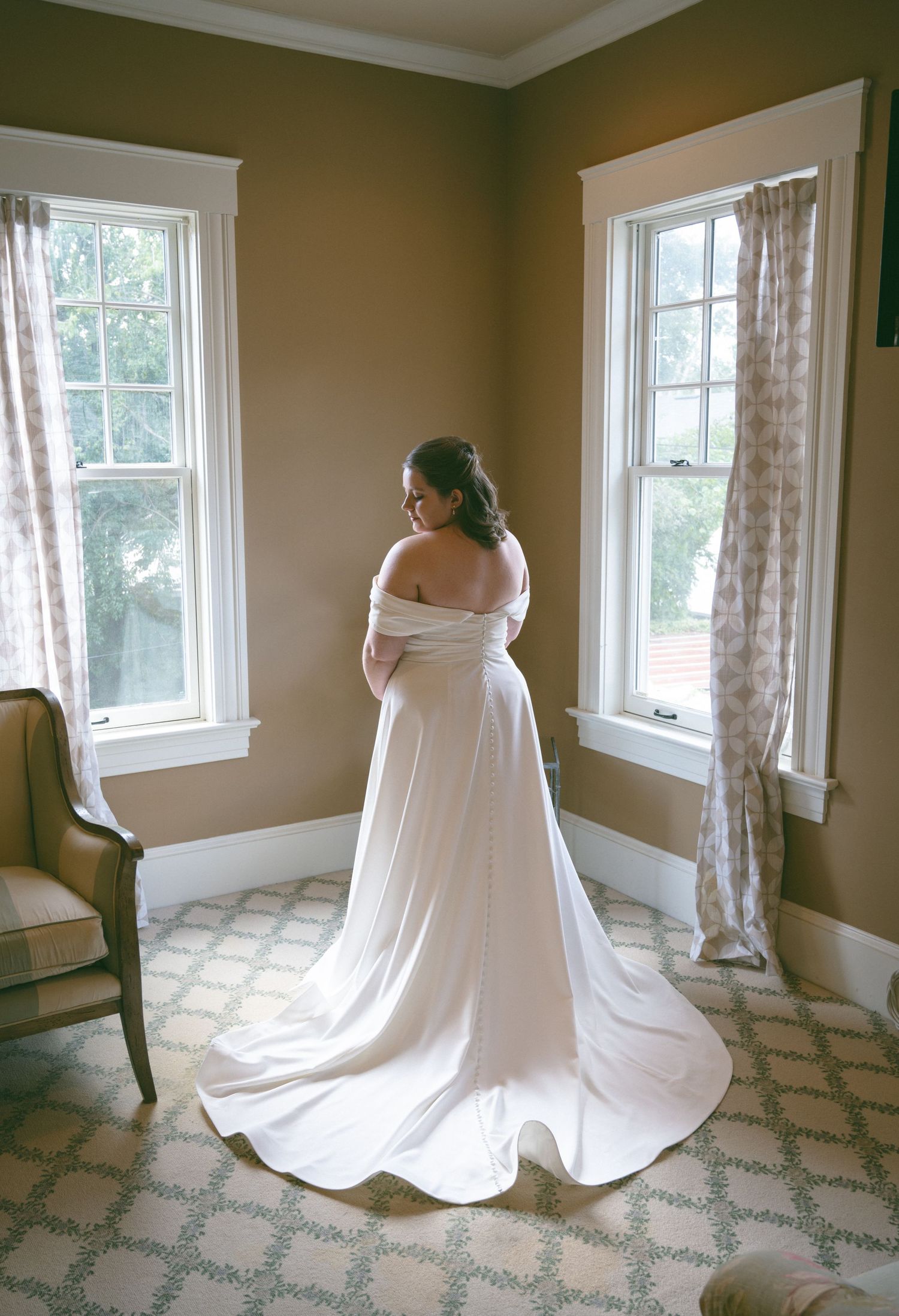 Bride in elegant off-shoulder white gown with long train stands between two windows in soft light.