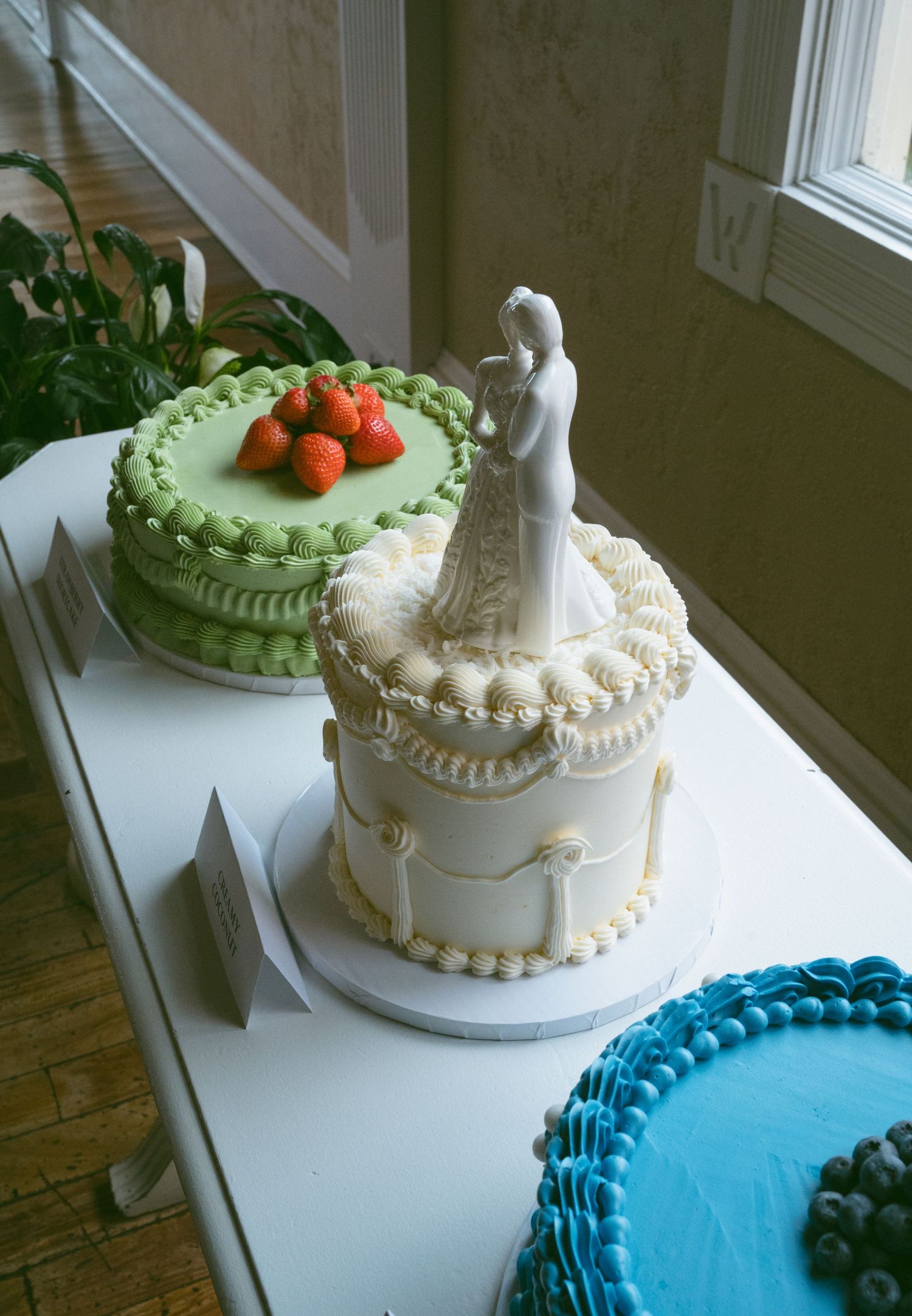 Elegant three-tier white wedding cake with bride and groom topper displayed alongside colorful cakes.