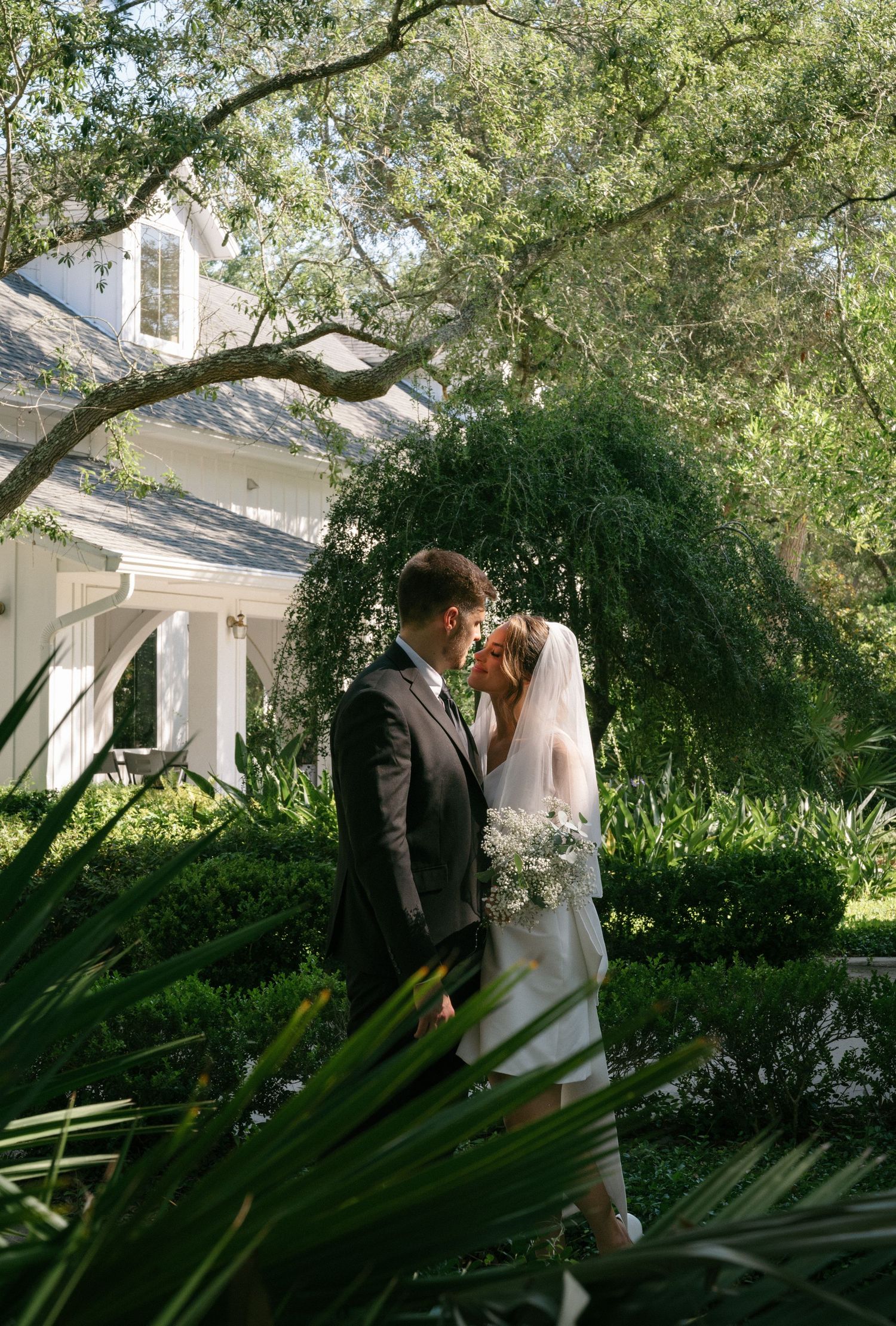 Bride and groom sharing intimate moment in lush green garden beside elegant white estate building.