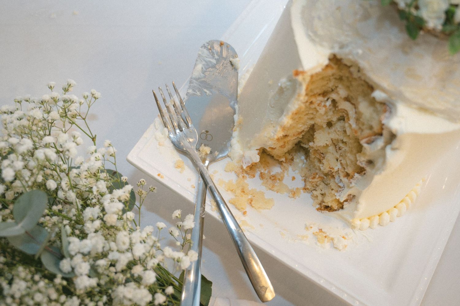 Partially cut wedding cake beside silver fork and knife server with baby's breath floral arrangement.
