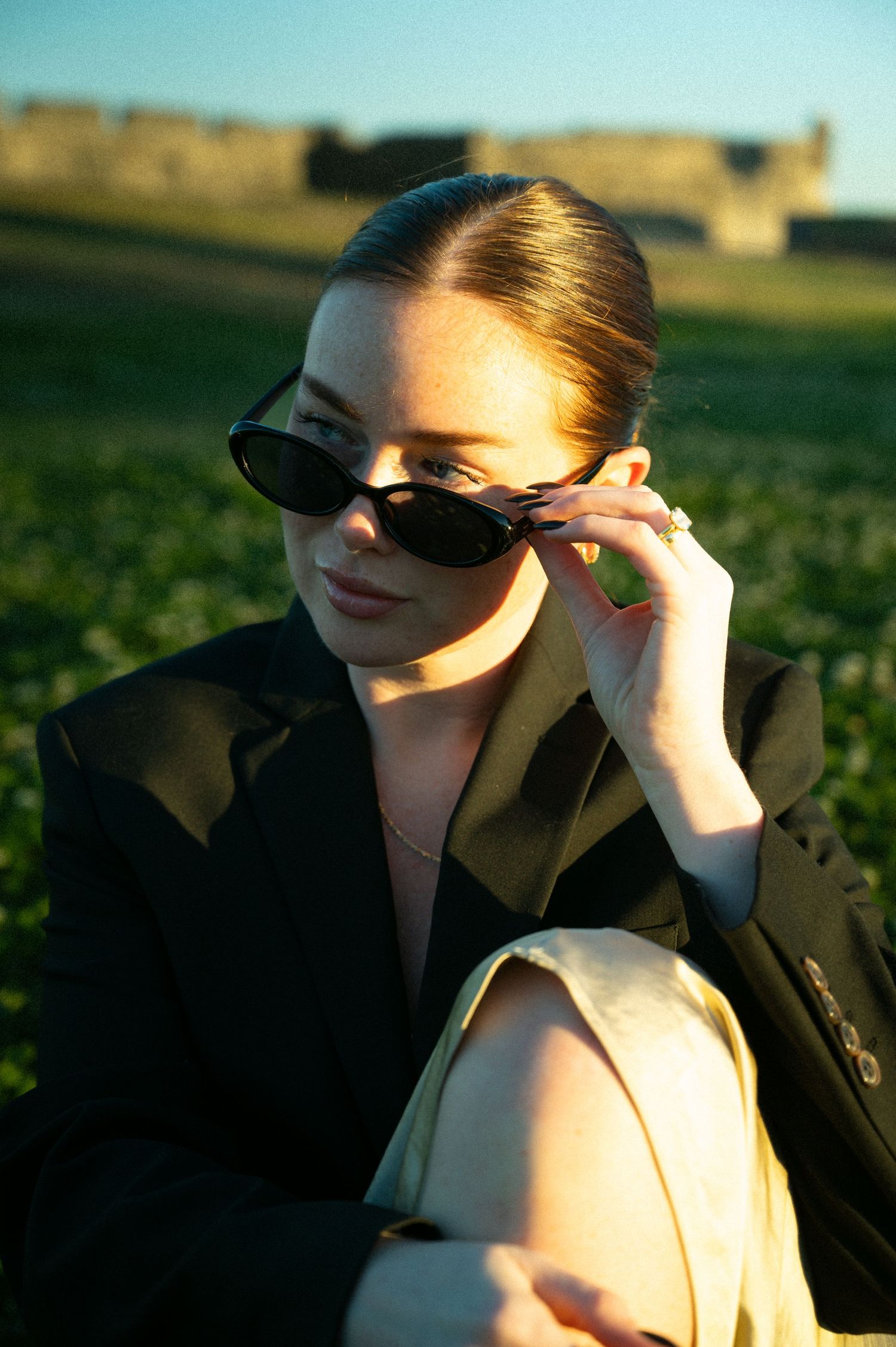 Chic woman in black blazer adjusting cat-eye sunglasses while sitting in a sunlit green field in Saint Augustine, Florida.