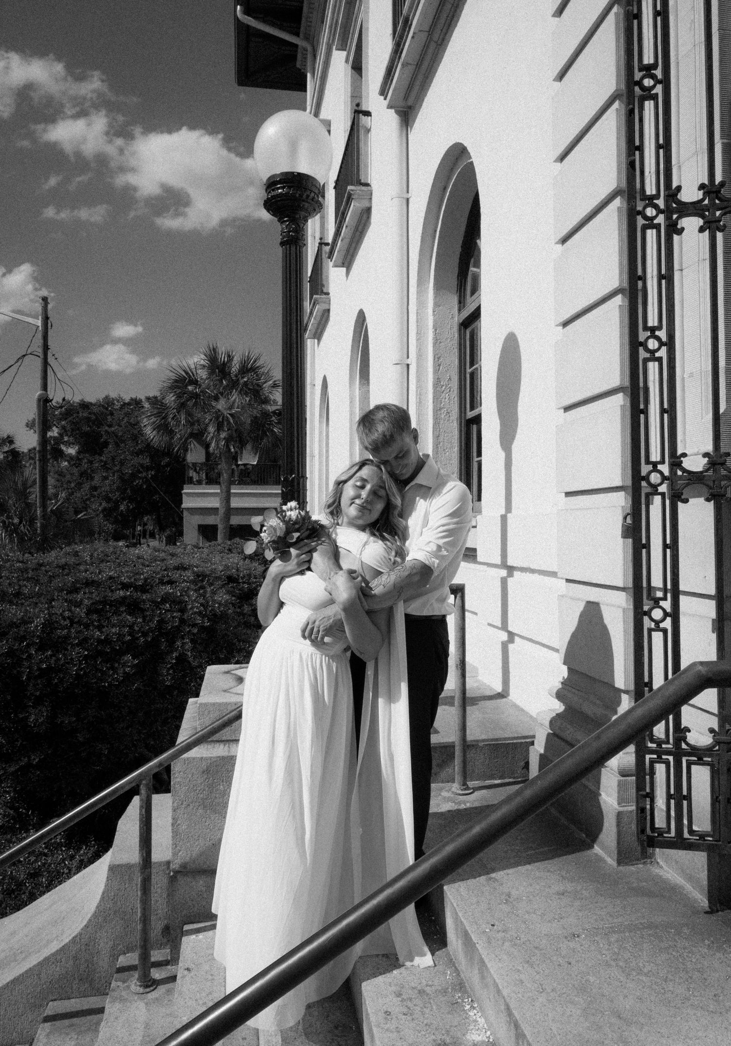 Black and white photo of groom embracing bride with bouquet on steps of classic white building on Amelia Island, Florida.