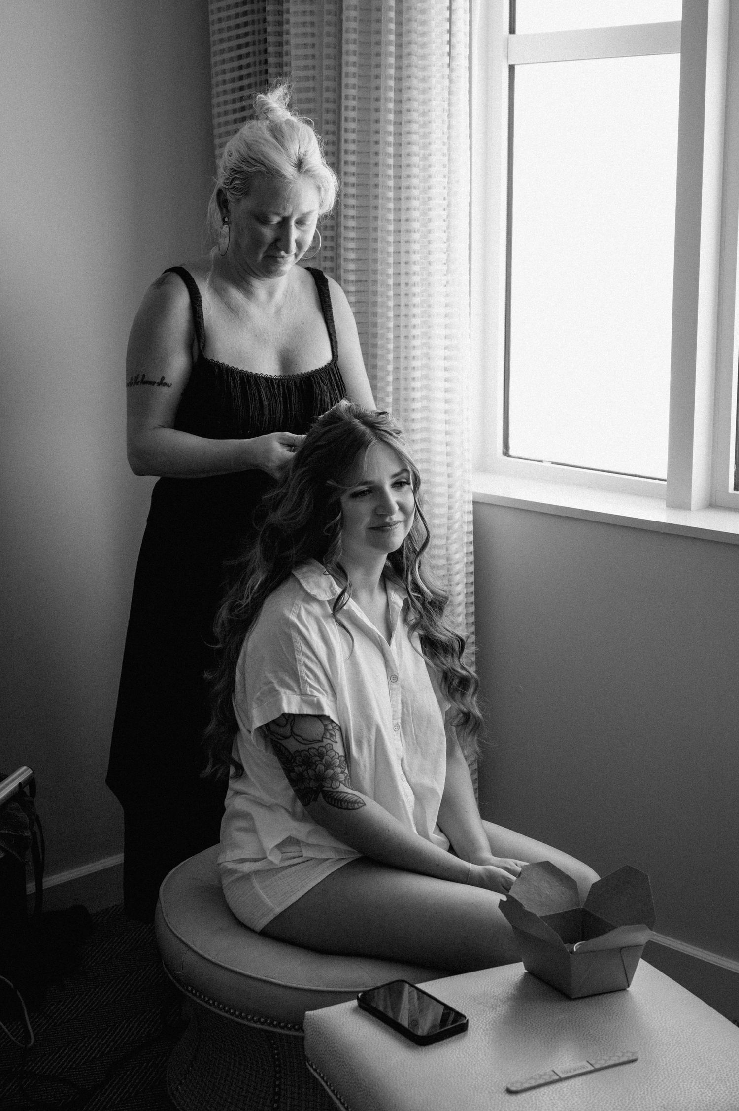 Black and white photo of bride getting hair styled by hairdresser near bright window.