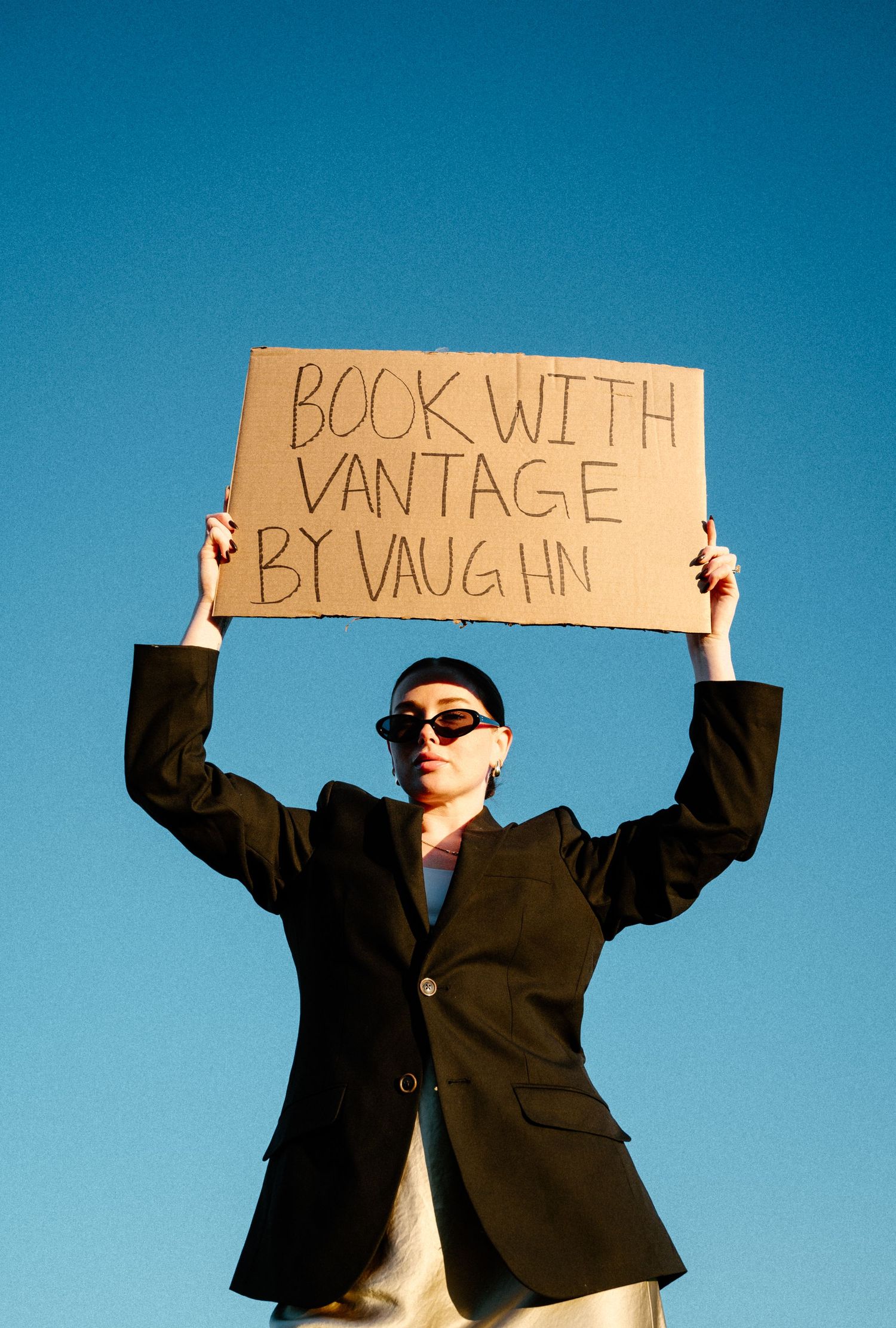 Woman in black blazer holds cardboard sign reading Book With Vantage By Vaughn overhead against blue sky.