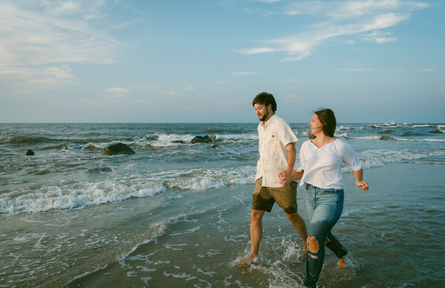 Couple walking hand in hand along ocean shoreline with waves and blue sky behind them.