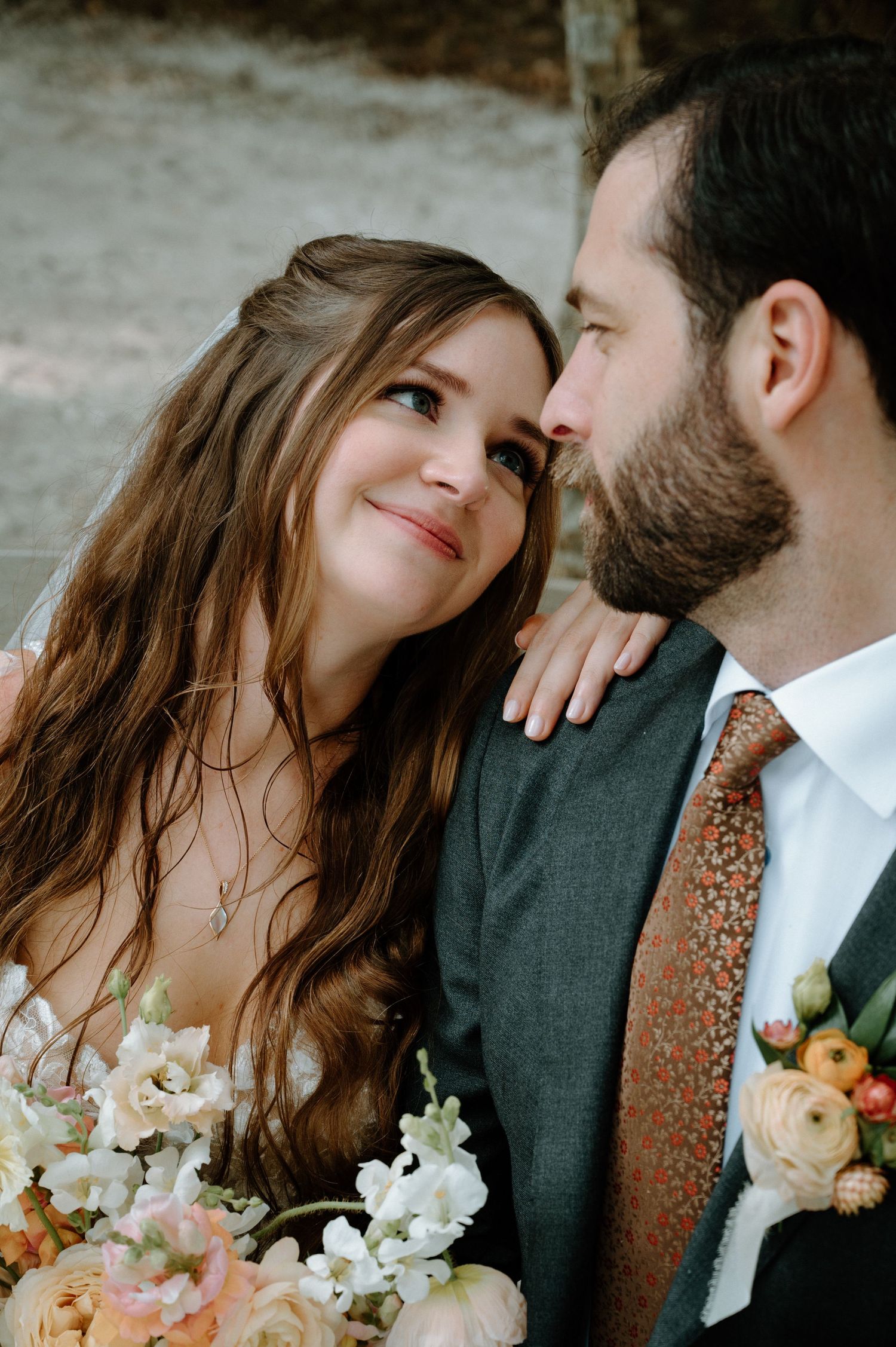 Bride gazes lovingly at bearded groom while holding peach and white floral bouquet.