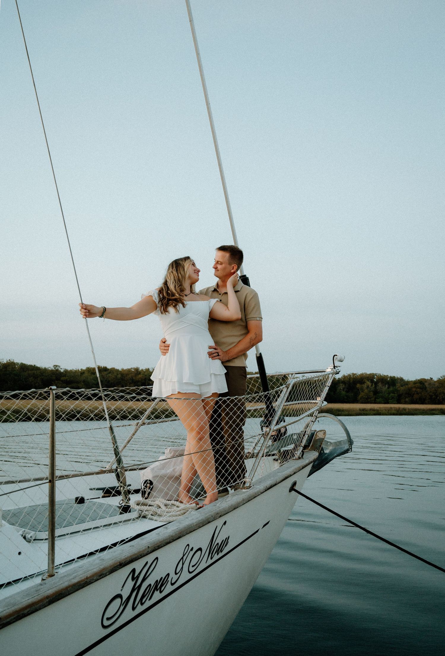 Couple embracing on sailboat named Here and Now at dusk on calm water.