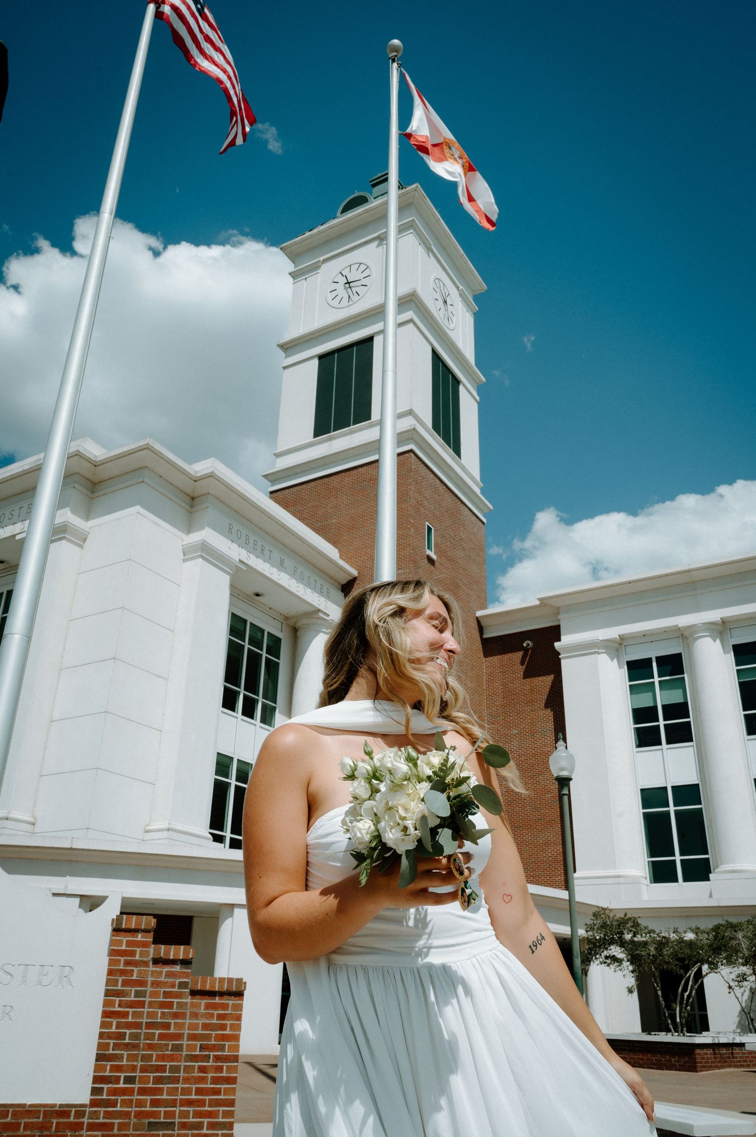 Bride in white dress holds bouquet outside brick courthouse with American and state flags.