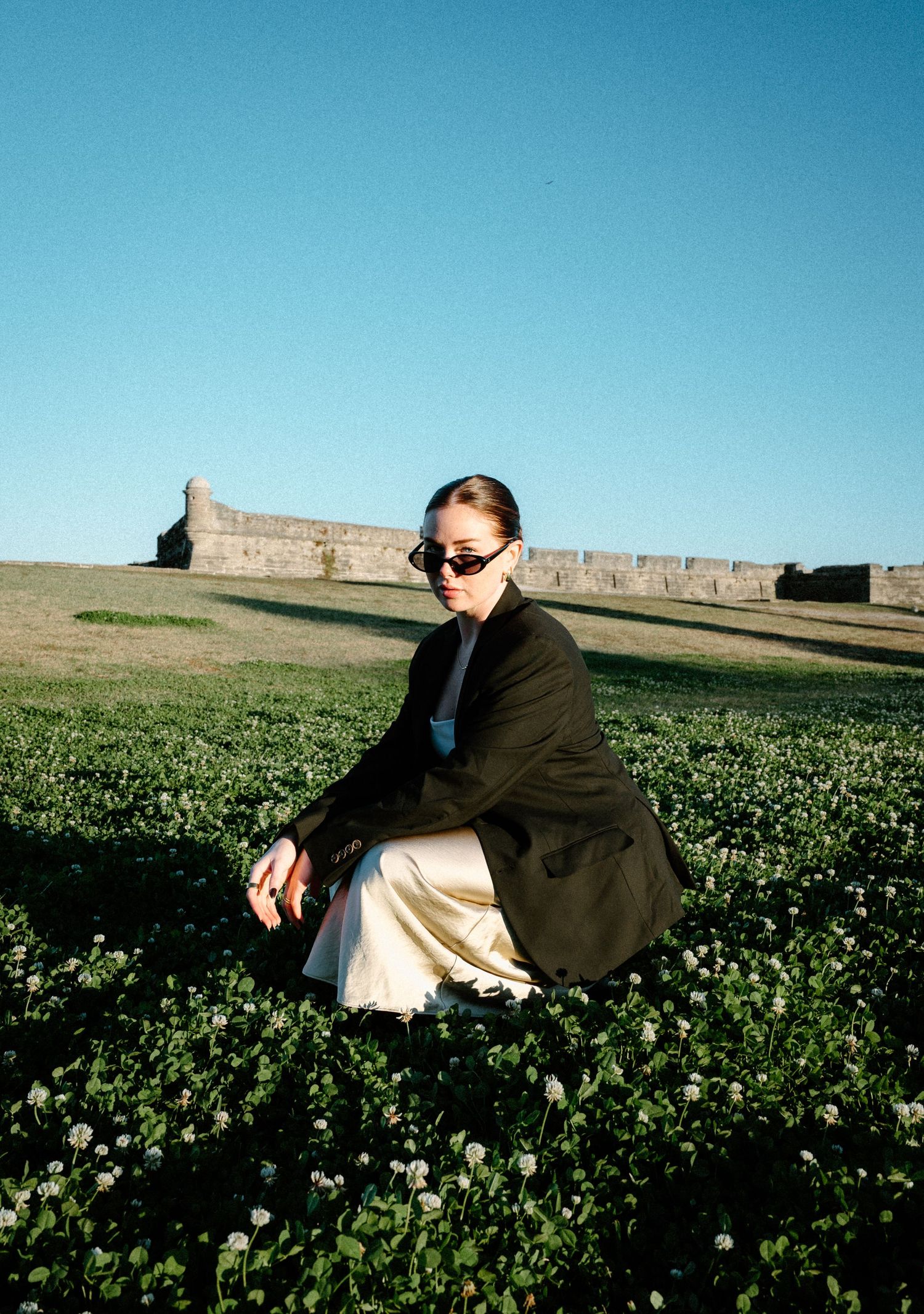 Woman in black blazer and gold skirt crouching in green field with historic stone fort ruins behind her in Saint Augustine, Florida.