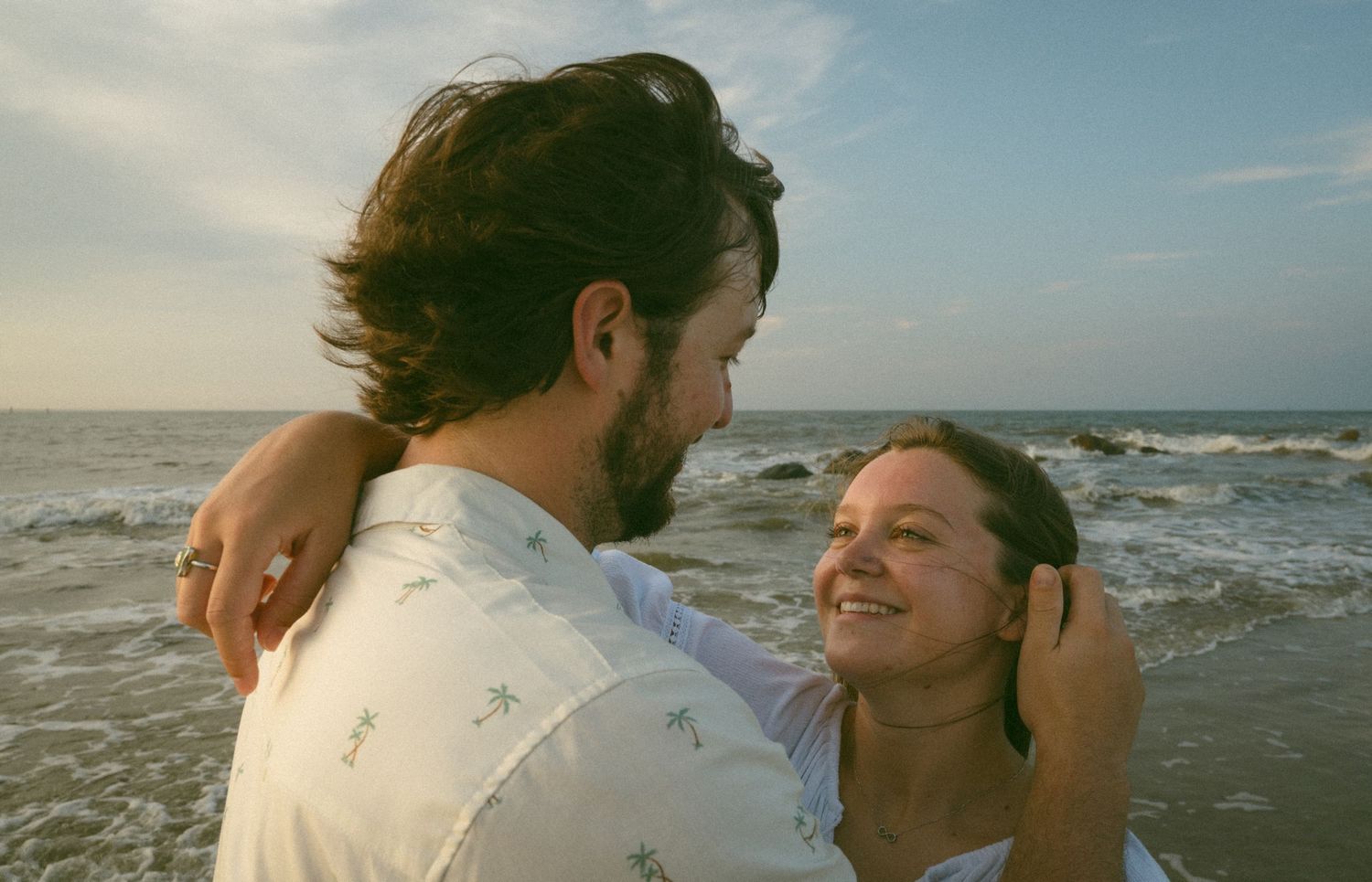 Couple smiling and embracing each other on a beach with ocean waves and sunset sky behind them.
