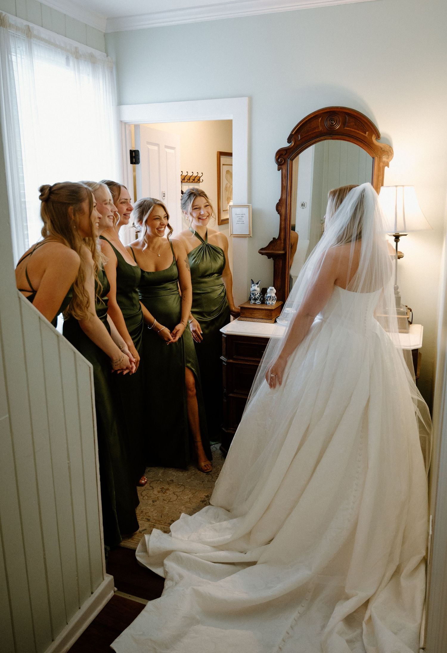 Bridesmaids in olive green dresses reacting with joy as bride reveals her wedding gown.