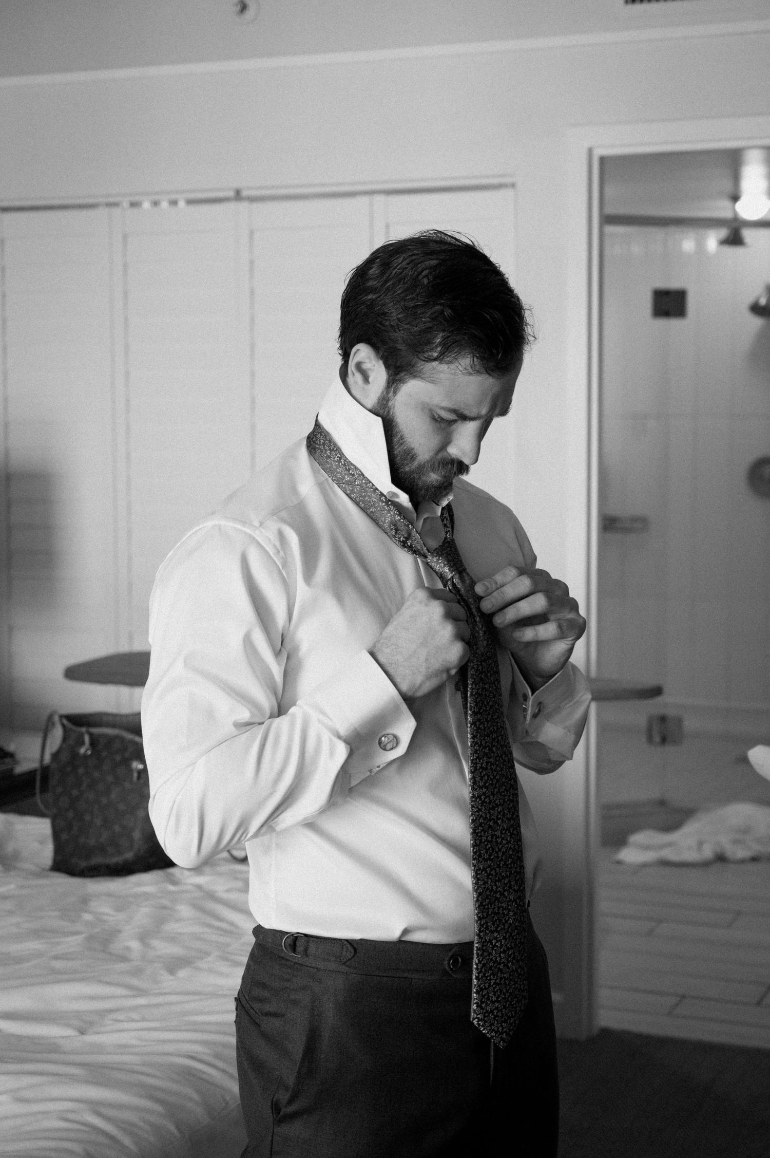 Groom adjusts patterned tie while getting ready in hotel room on wedding day.