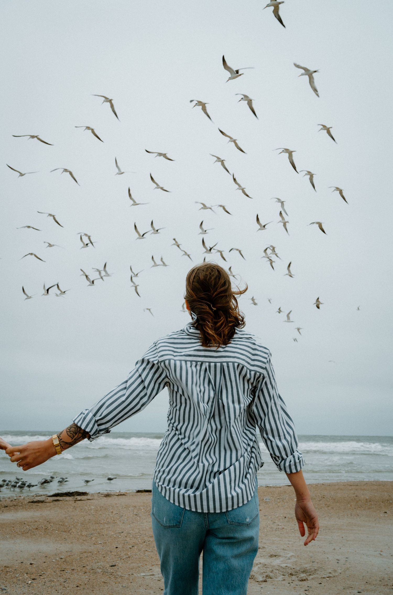 Woman in striped shirt stands on beach with arms out as seagulls fly overhead.