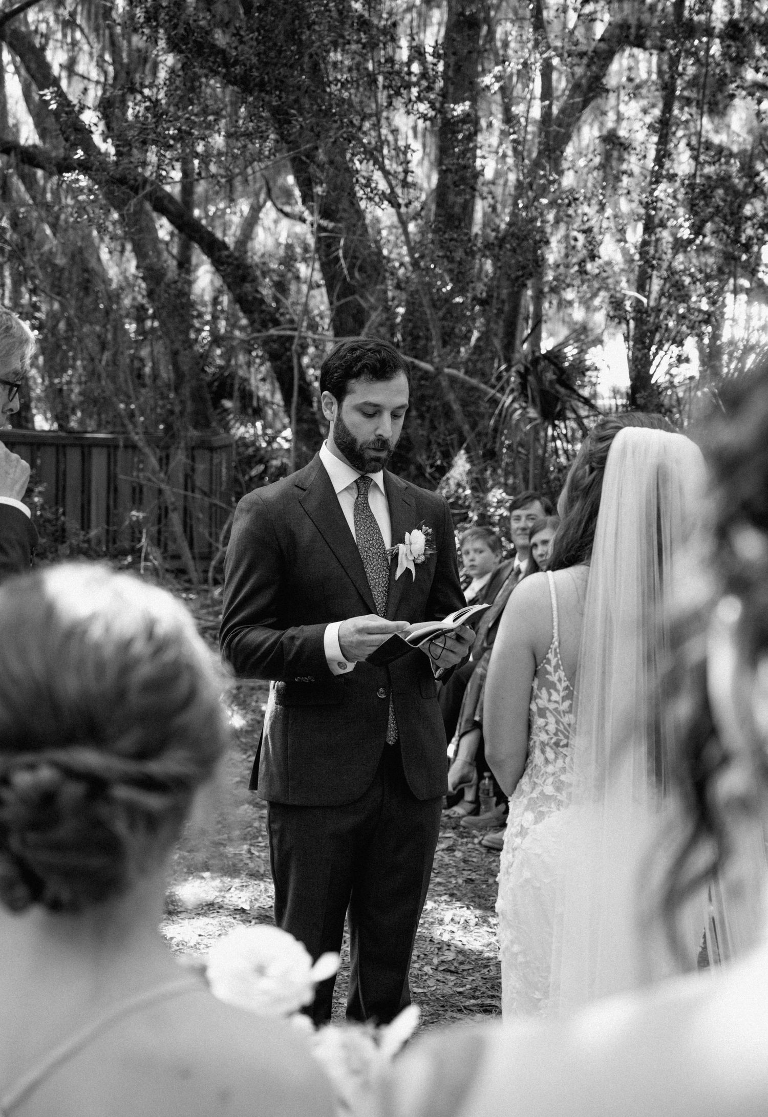 Groom reads vows from paper during outdoor wedding ceremony under moss-draped trees.