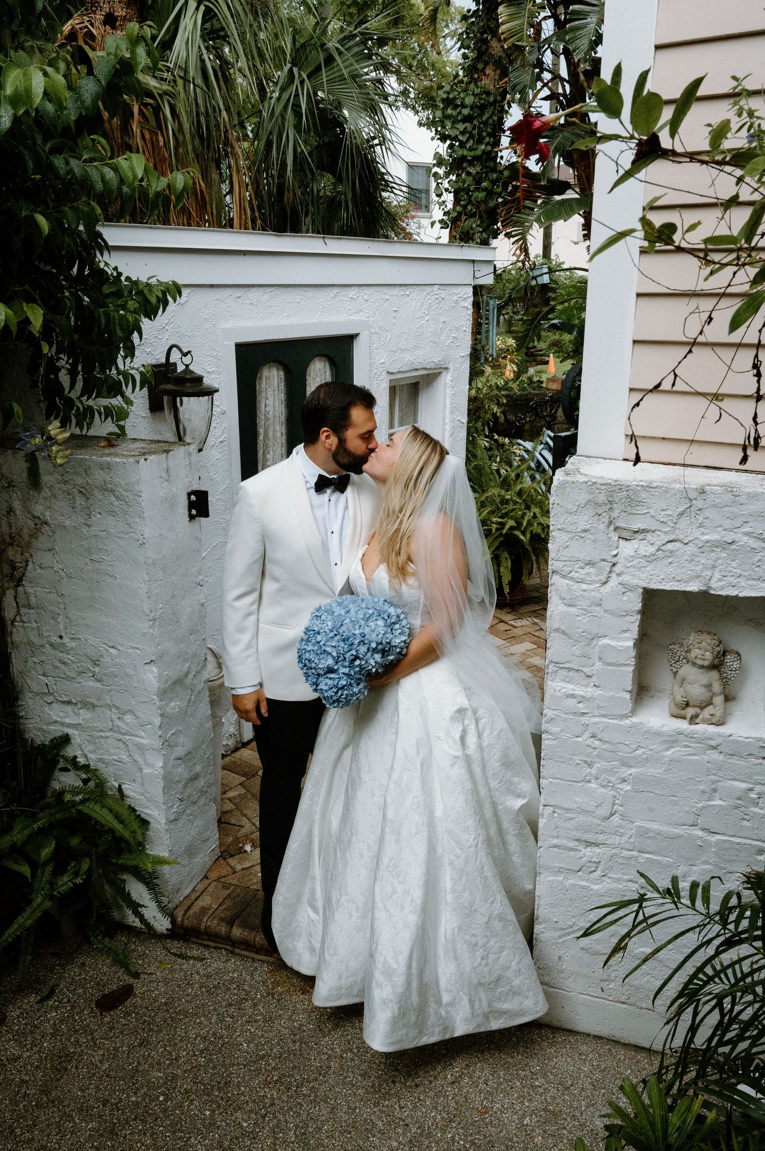 Bride and groom sharing first kiss outdoors surrounded by lush greenery with blue hydrangea bouquet.