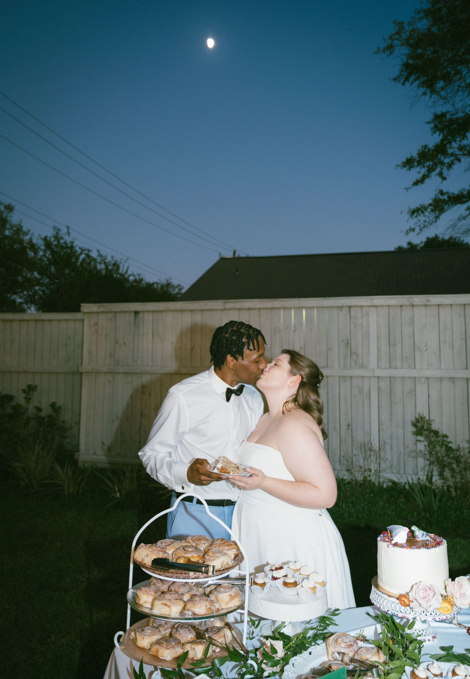 Couple shares a kiss at outdoor wedding dessert table under a moonlit night sky.