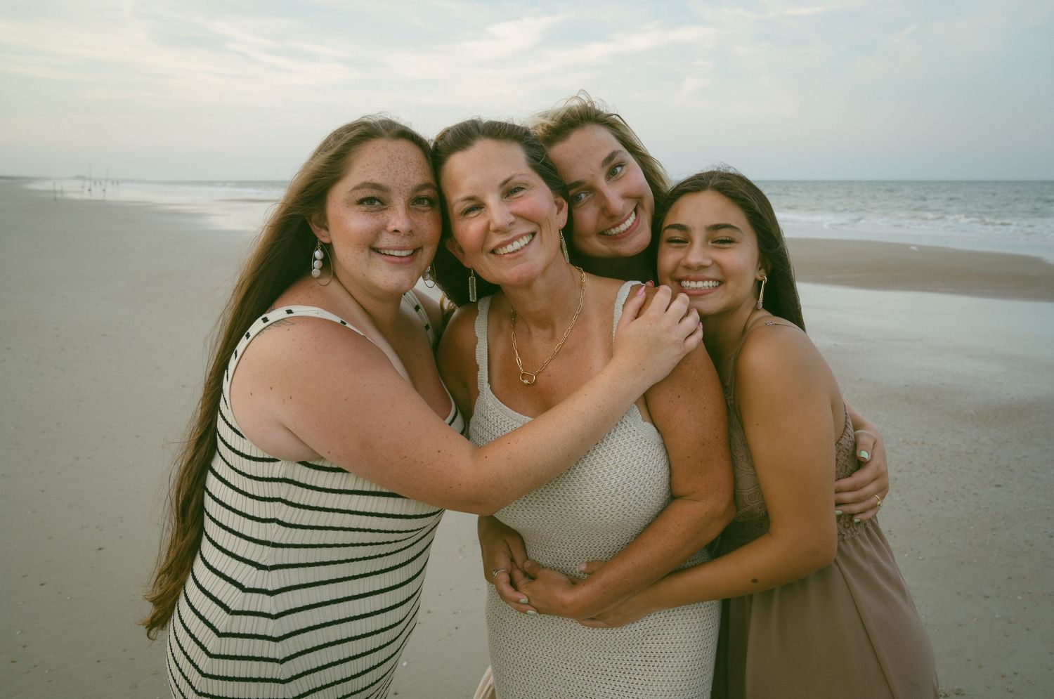 Four women smiling and embracing on a beach at dusk with ocean waves behind them.
