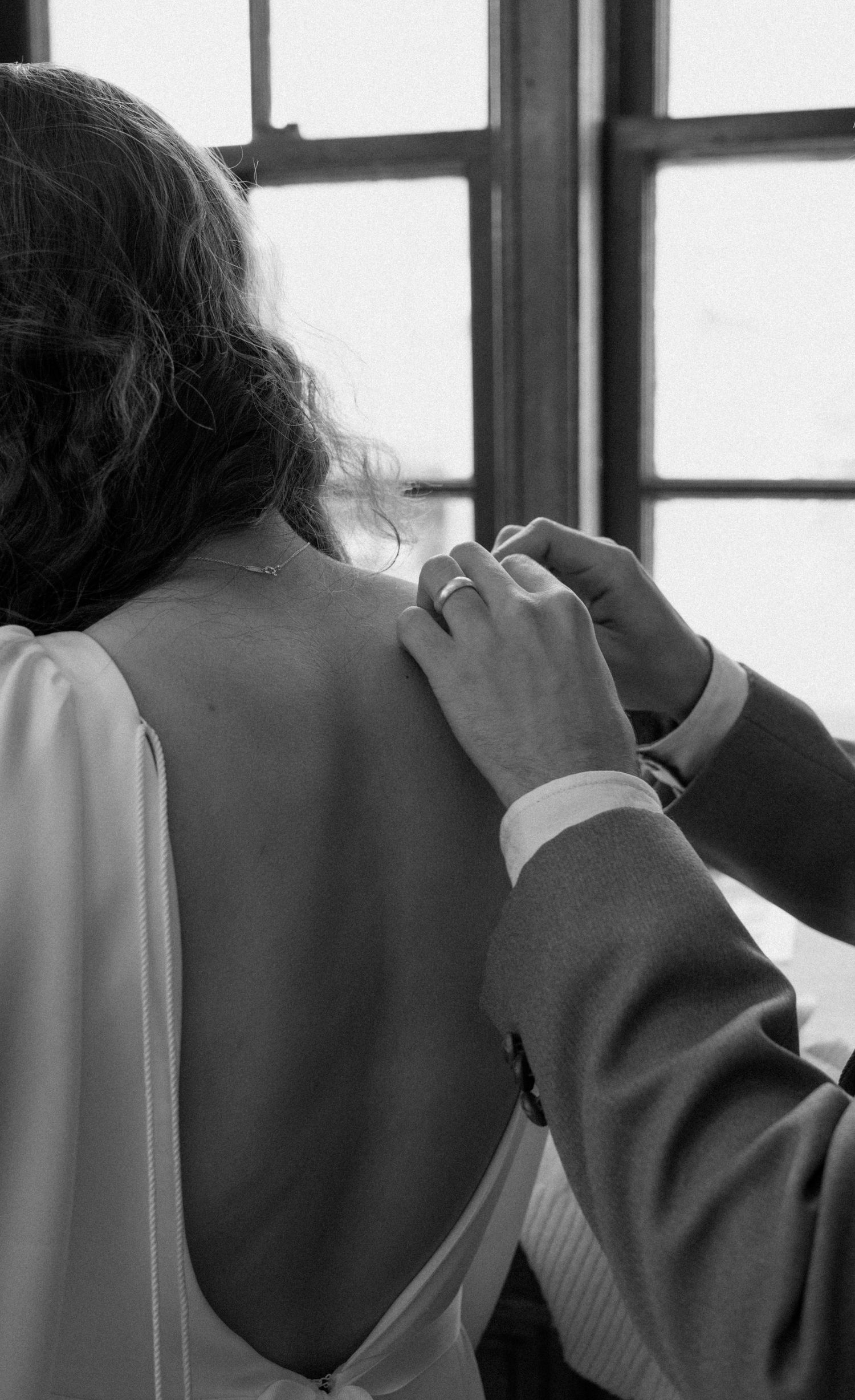 Black and white photo of someone fastening the back of a bride's white wedding dress near a window.