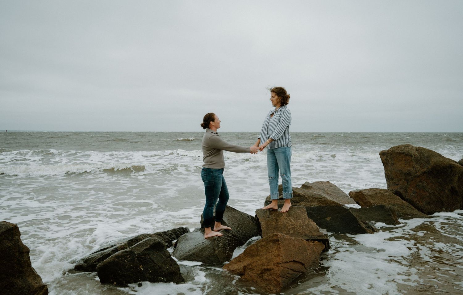 Two people holding hands while standing on rocky shore with waves crashing behind them on Amelia Island, Florida.