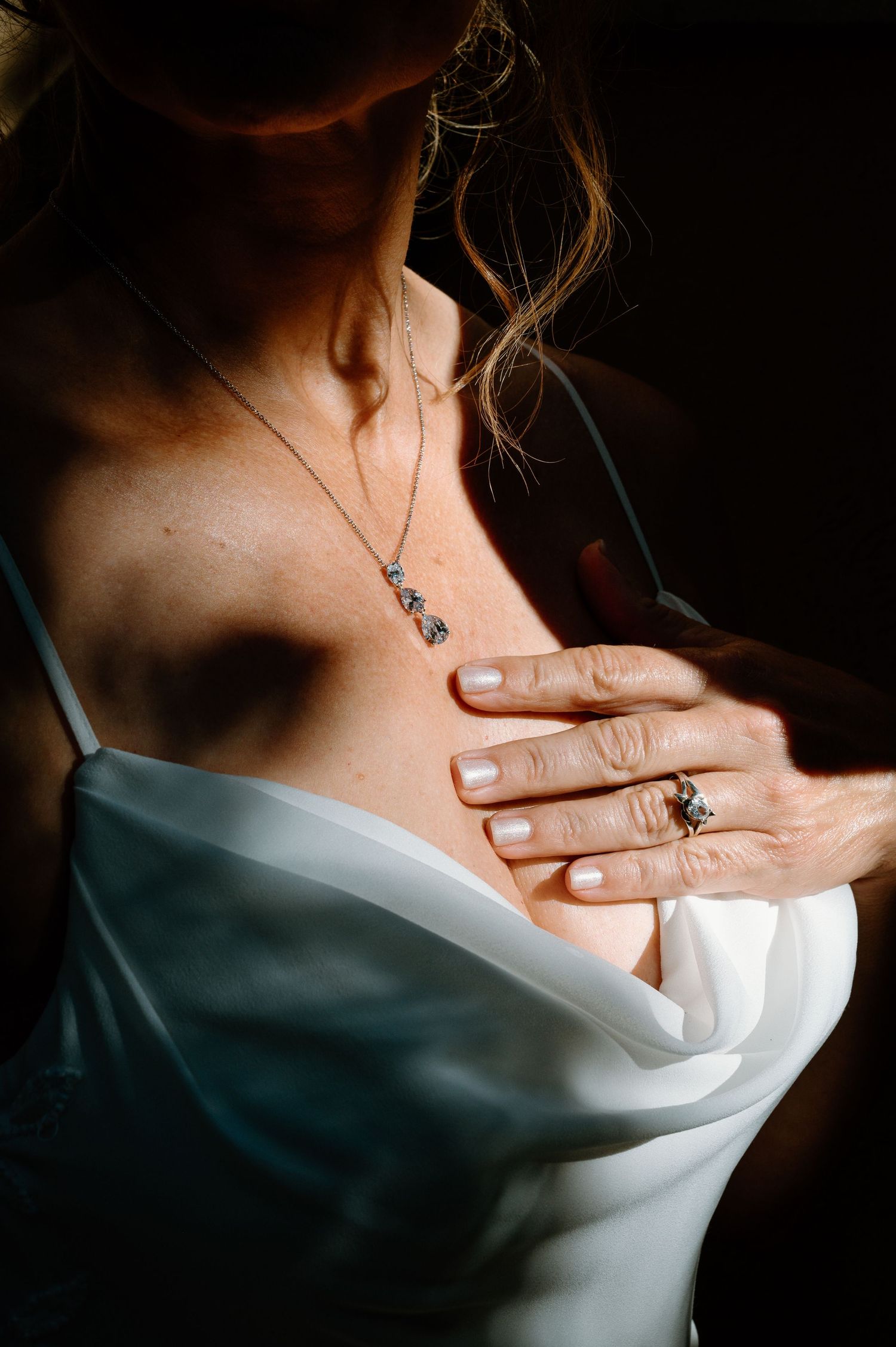 Close-up of bride in white gown touching her chest, wearing a delicate necklace and ring.
