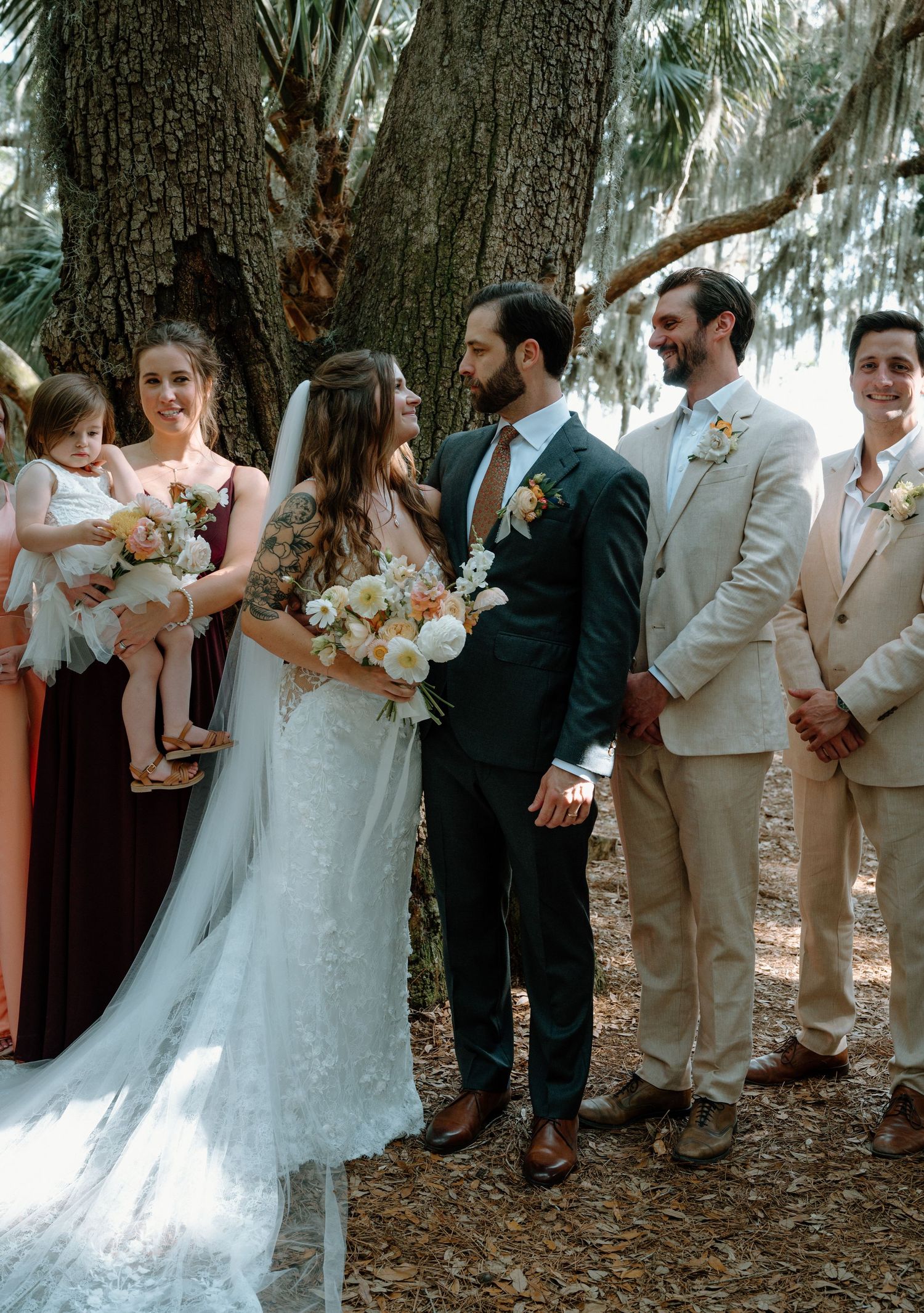 Bride and groom share a smile with wedding party beside a large tree outdoors.