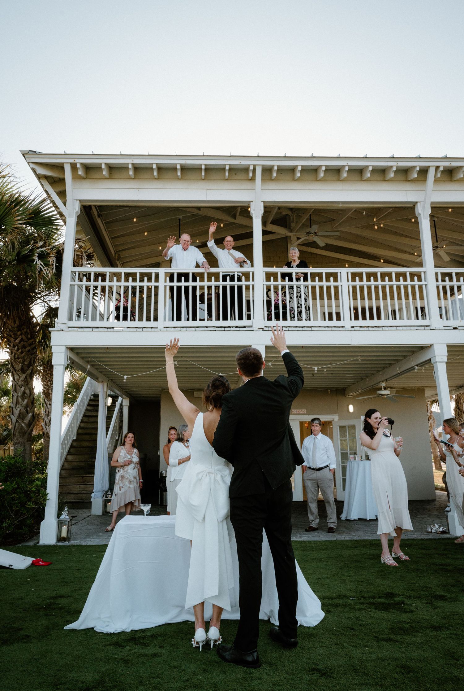 Newlyweds raise hands toward cheering guests on balcony during outdoor wedding reception celebration on Amelia Island, Florida.