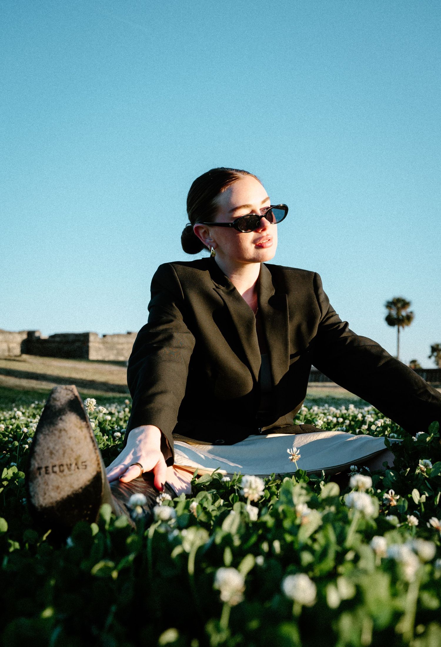 Woman in black blazer and sunglasses poses among white wildflowers near historic stone fort on clear day.
