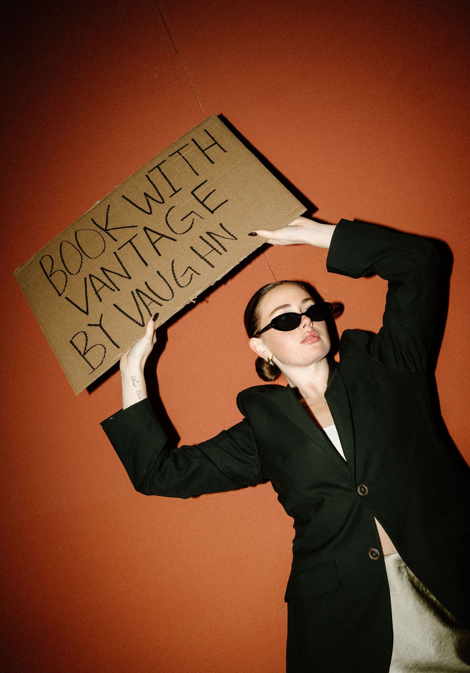 Woman in black blazer holds cardboard sign reading Book With Vantage By Vaughn against orange wall.