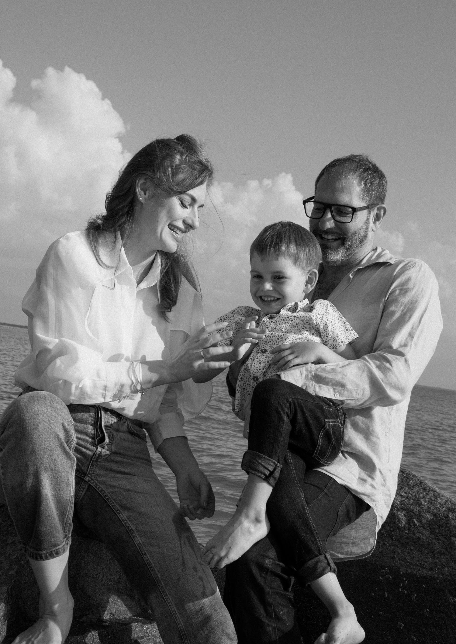 Black and white photo of a happy family with a toddler sitting by the ocean on a sunny day at Fort Clinch State Park on Amelia Island, FL.