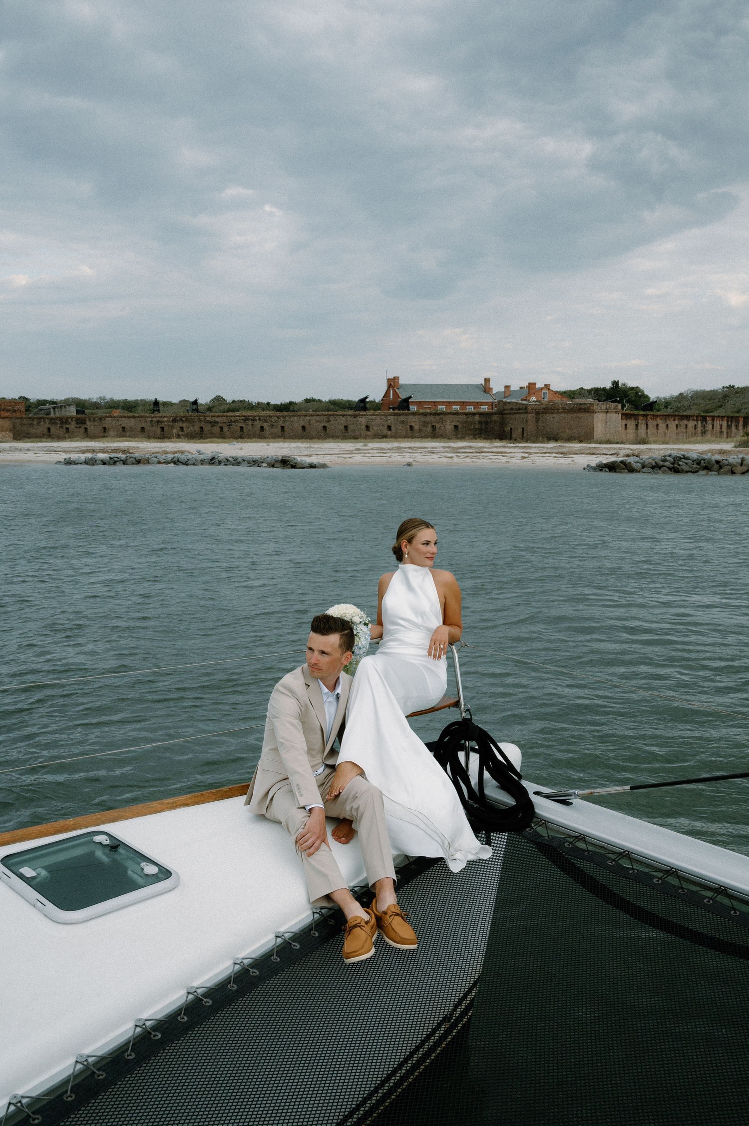 Bride and groom sitting on the bow of a boat on calm water with a historic fort in the background.