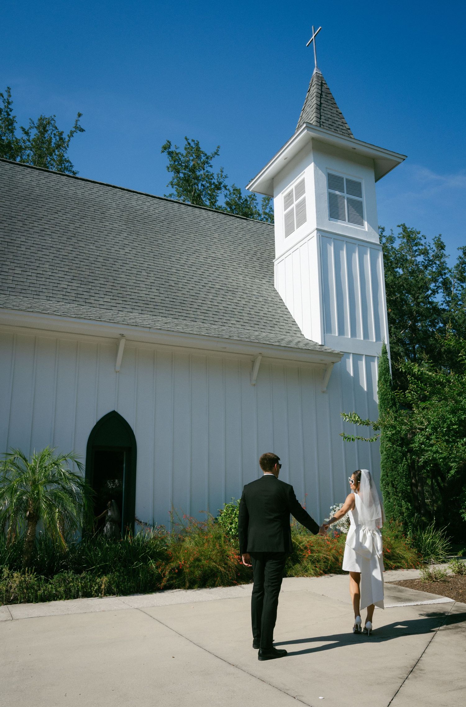 Bride and groom holding hands walking toward a white chapel with a cross on a sunny day.