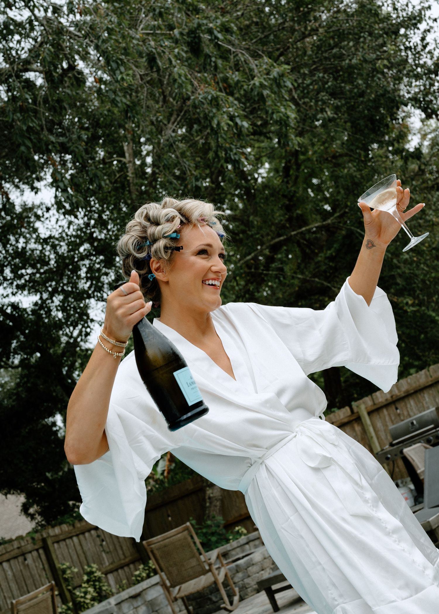 Joyful woman in white robe celebrating with champagne bottle and glass outdoors with hair in curlers.