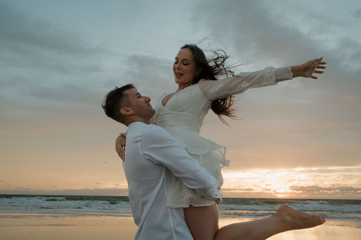 Groom lifting joyful bride on a windy beach at sunset, both dressed in white on Amelia Island, Florida.