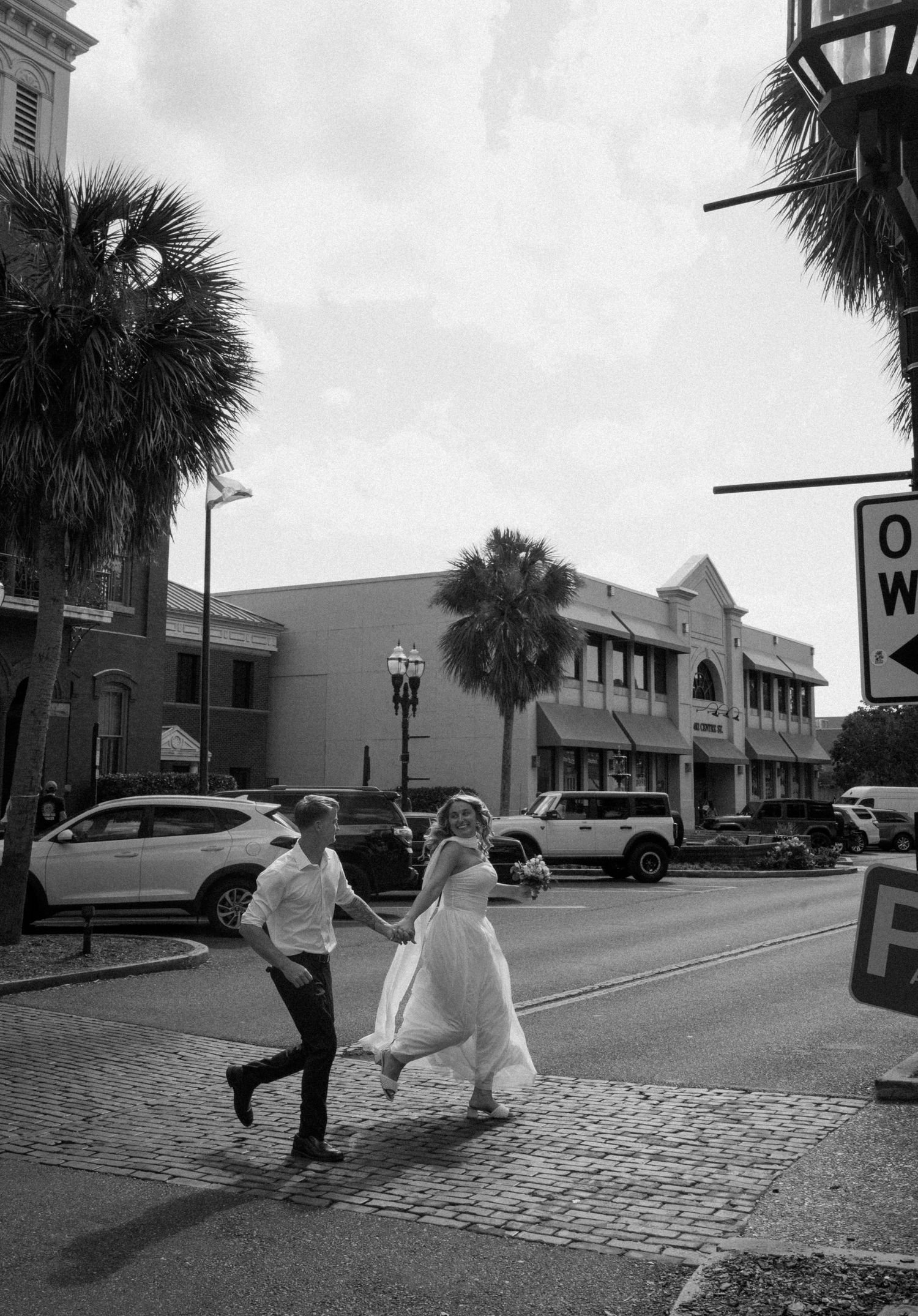 Newlywed couple running hand-in-hand across cobblestone street lined with palm trees in black and white in downtown Amelia Island.