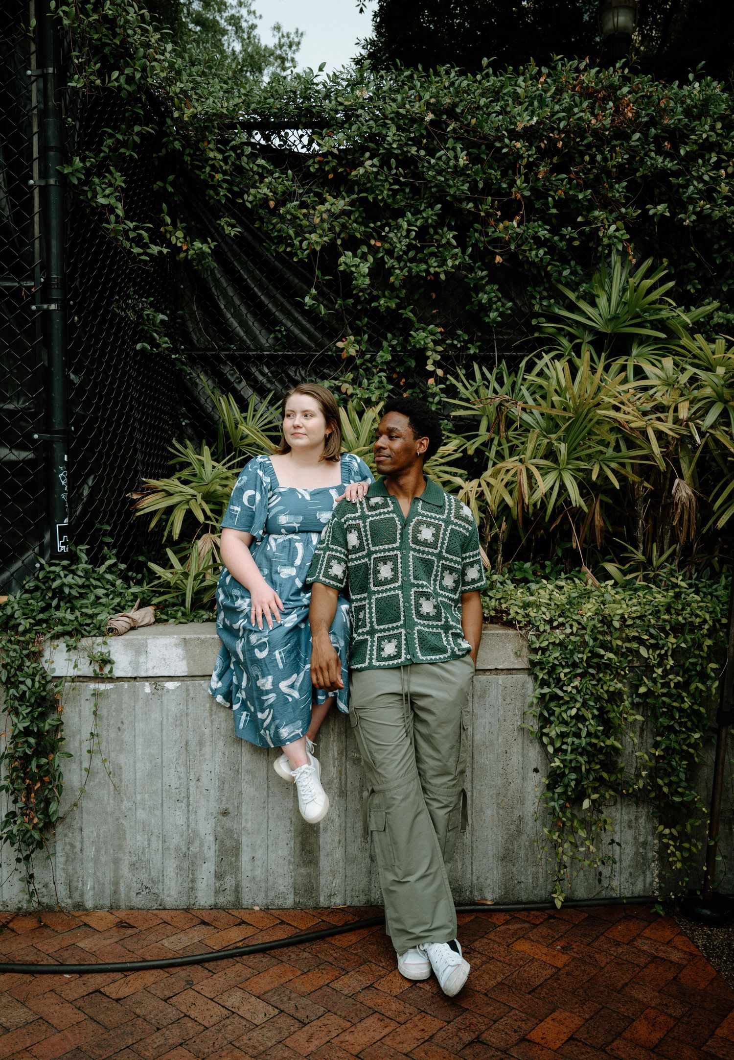 Couple leaning against a garden wall surrounded by lush green foliage on a brick path.