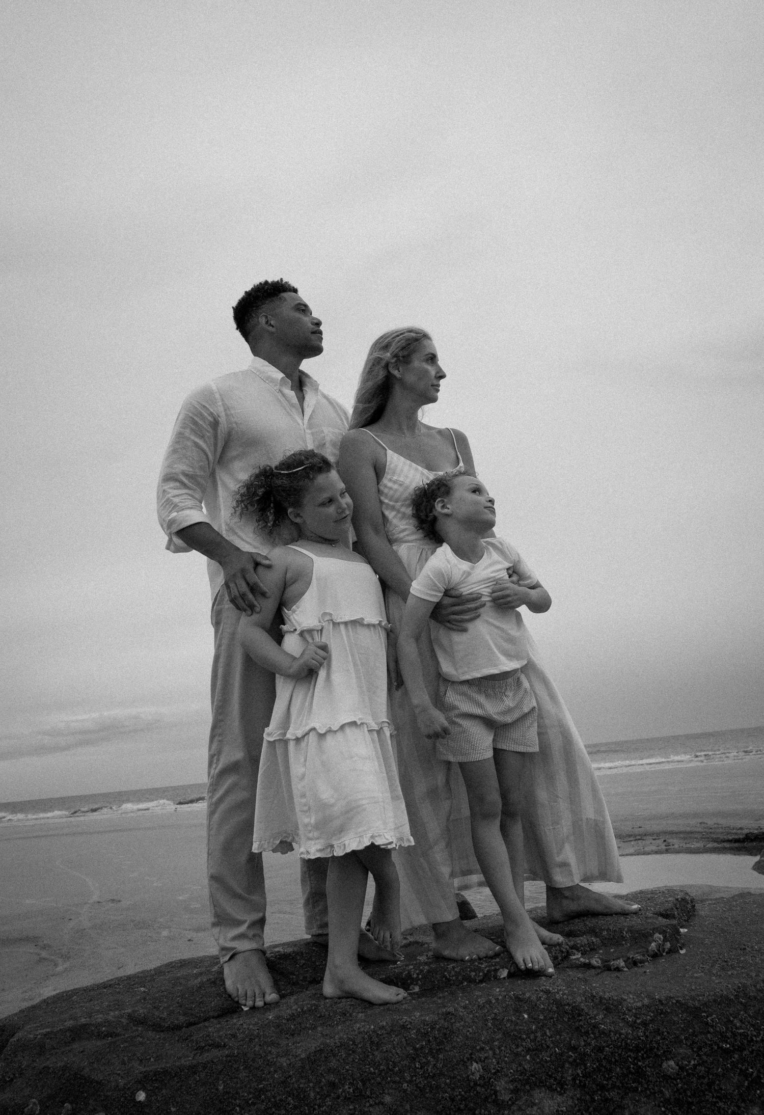 Black and white family portrait of parents with two children standing on rocks at the beach.