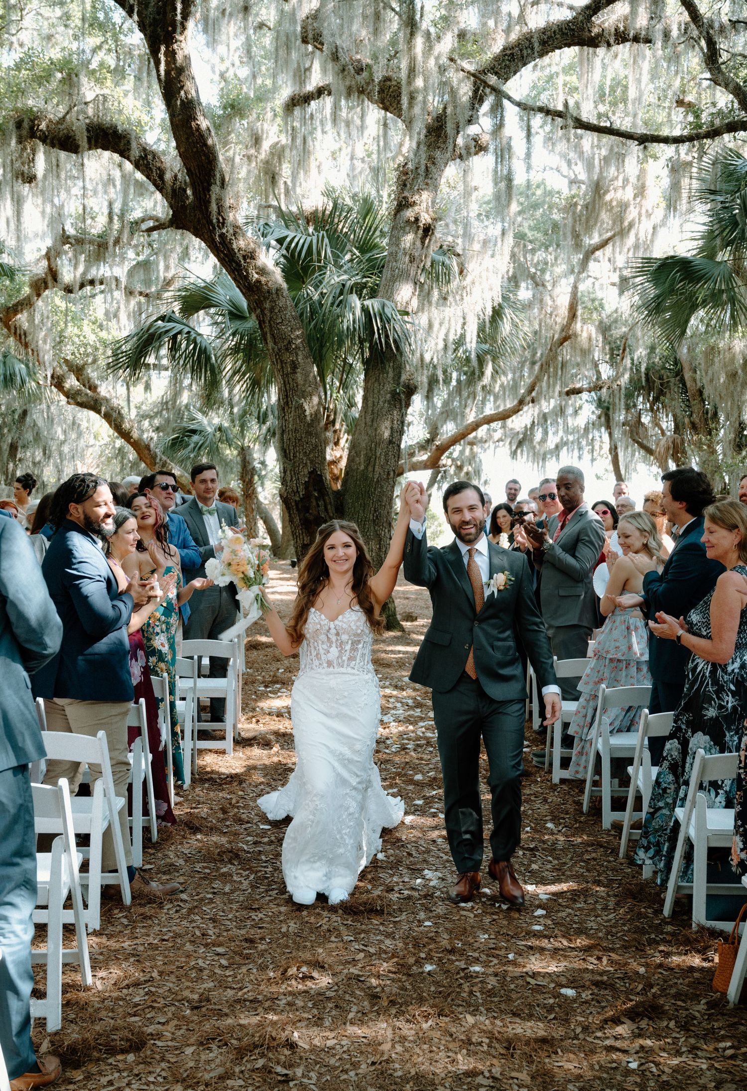 Joyful newlyweds walking back down aisle under Spanish moss-draped oak trees with cheering guests.