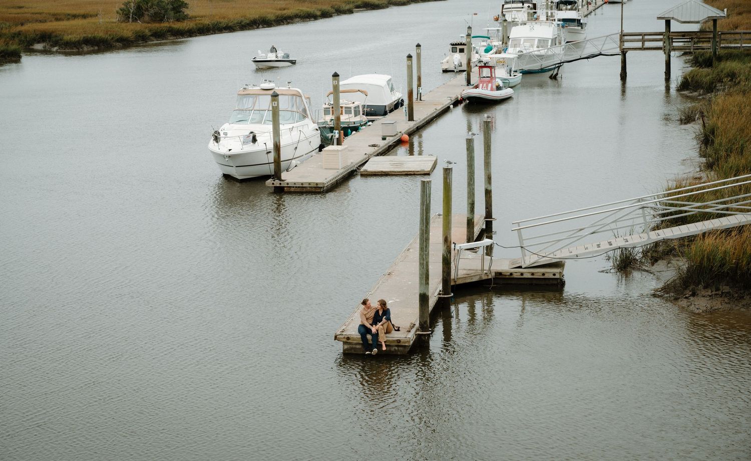 Aerial view of a couple sitting together on a wooden dock surrounded by boats.