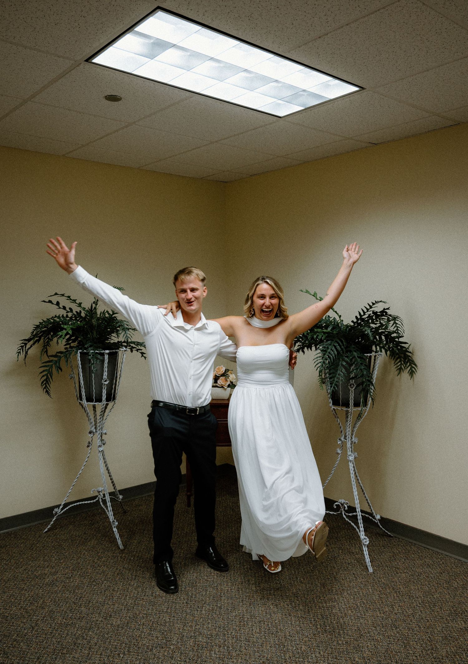 Jubilant newlyweds raise arms in celebration after courthouse elopement ceremony in small office room.
