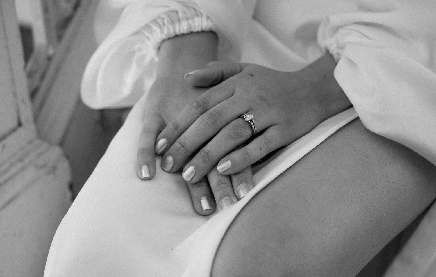 Black and white close-up of hands with a diamond engagement ring resting on a white dress.