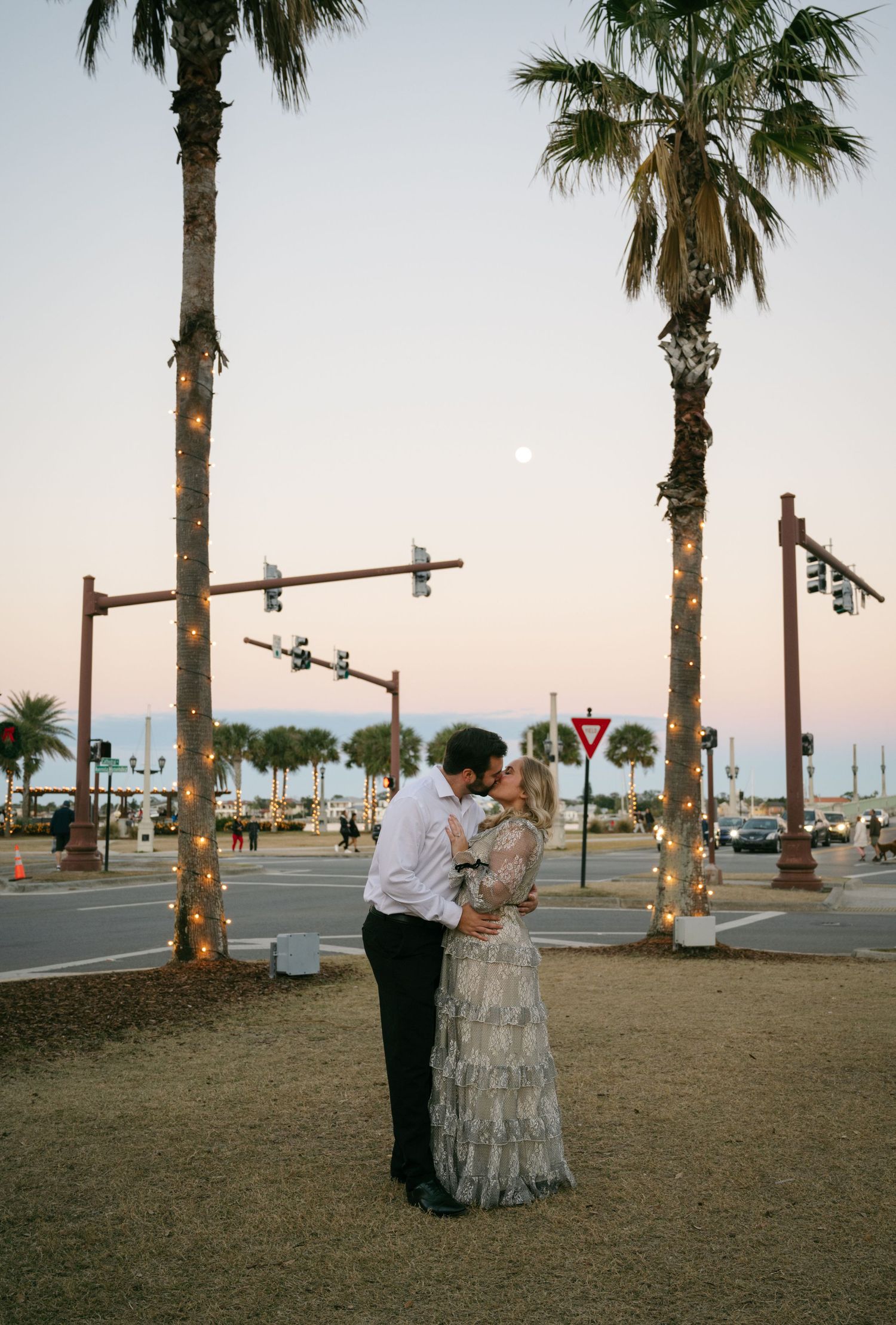 Couple sharing a kiss at sunset beneath palm trees wrapped in lights at a street corner.