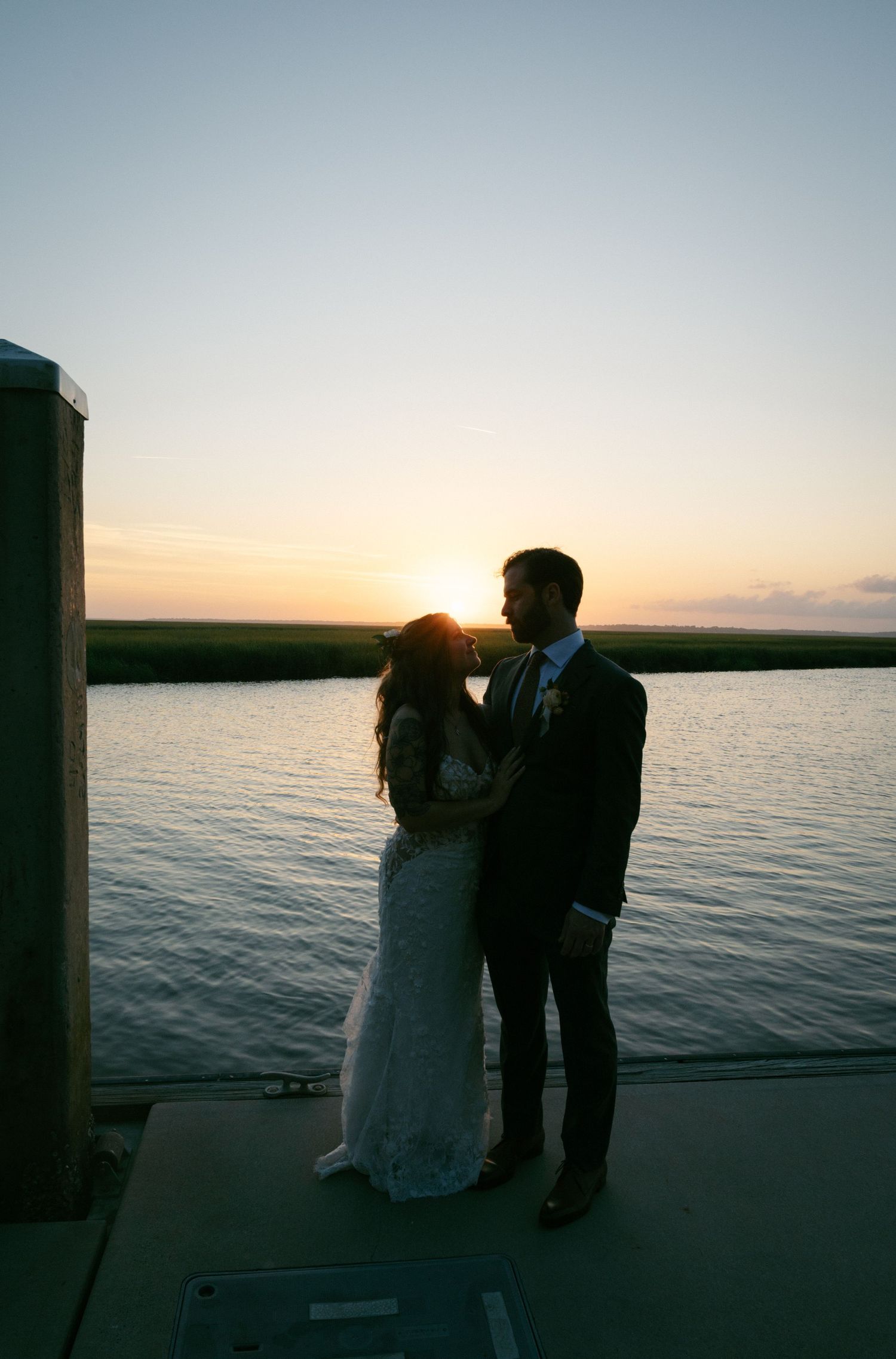 Bride and groom silhouetted against golden sunset over marsh waterway on dock.