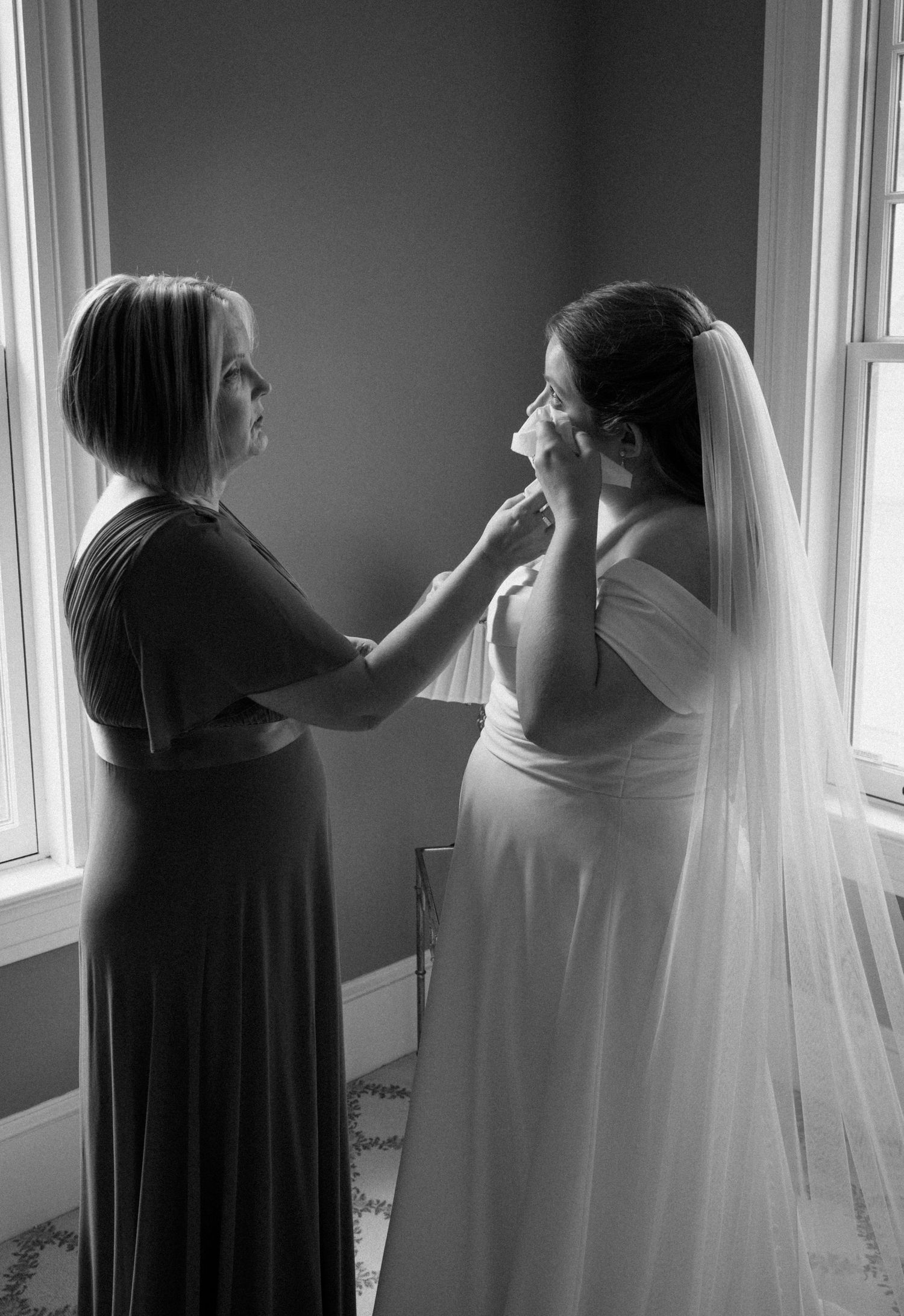 Black and white photo of bridesmaid tenderly wiping tears from emotional bride's face before wedding ceremony.