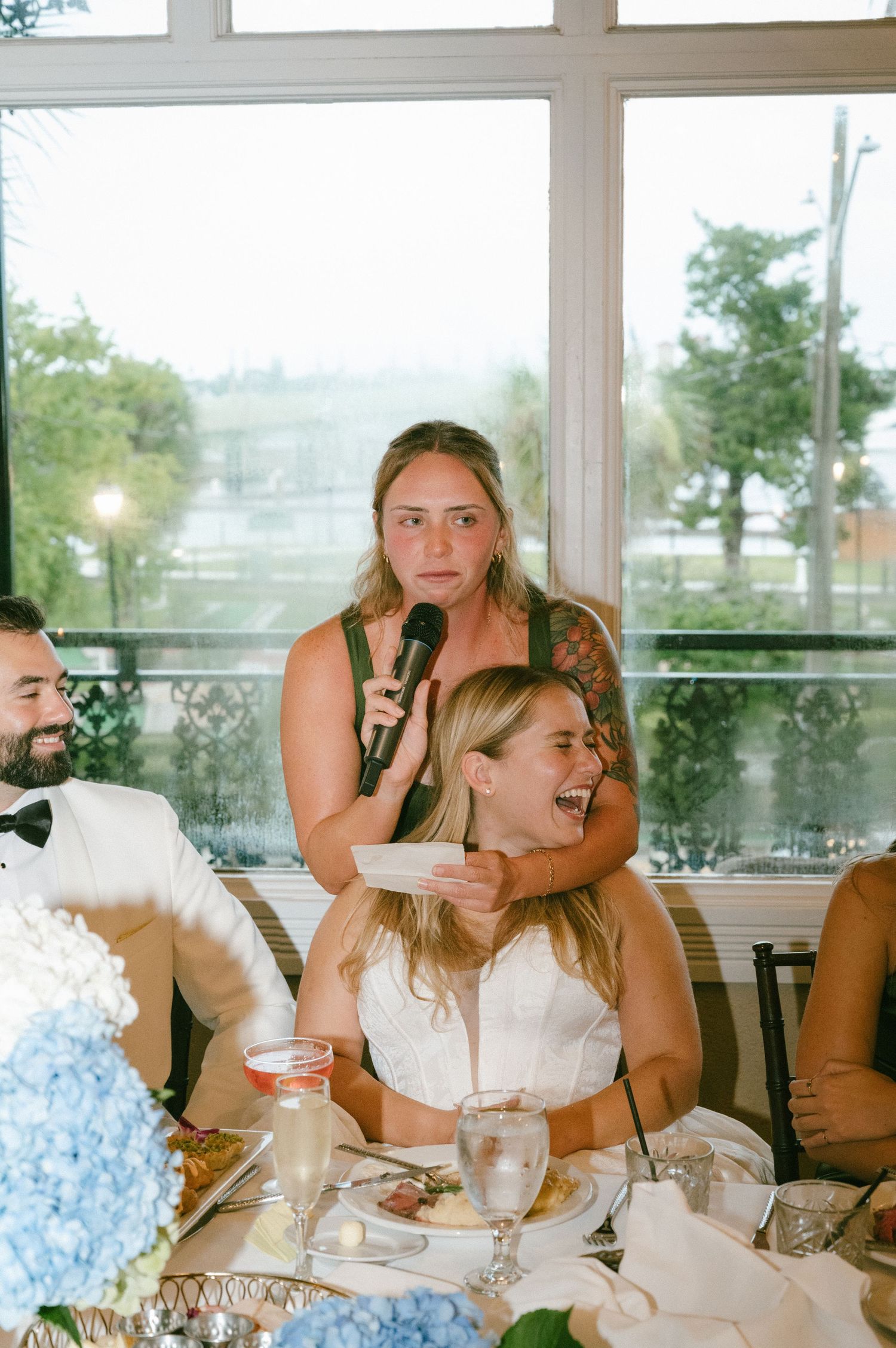 Bridesmaid giving heartfelt speech at wedding reception while bride laughs joyfully at table.