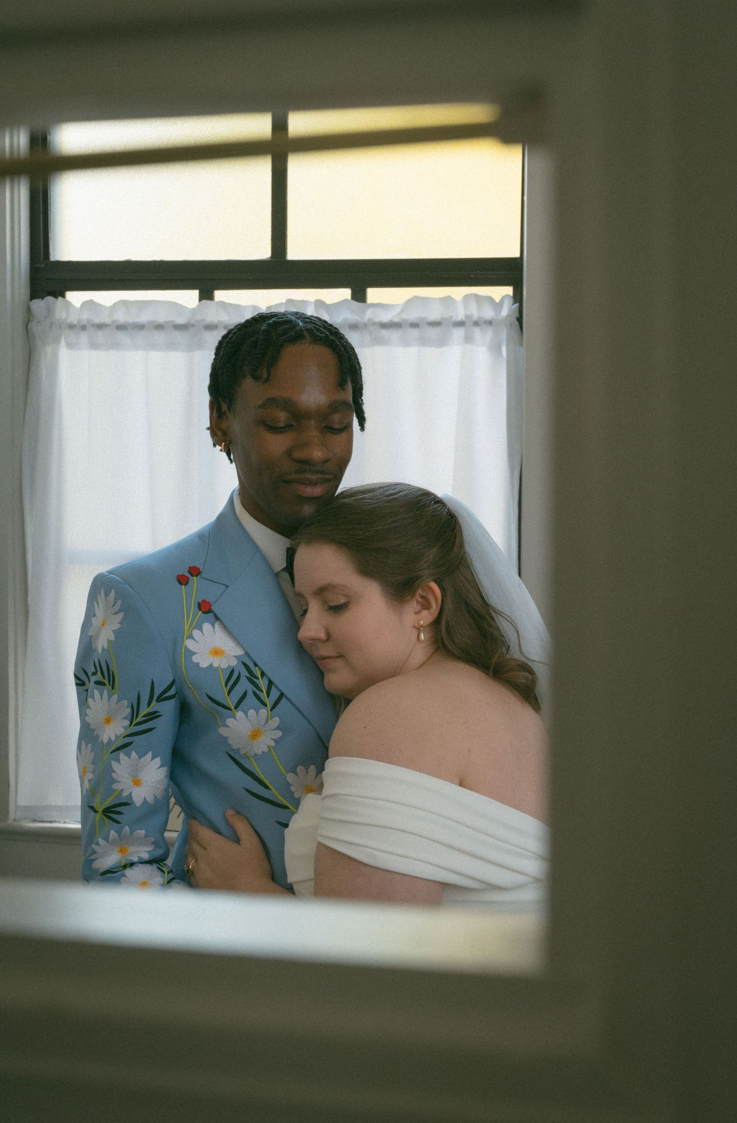 Interracial couple embracing in wedding attire, groom in floral blue suit and bride in white off-shoulder gown.