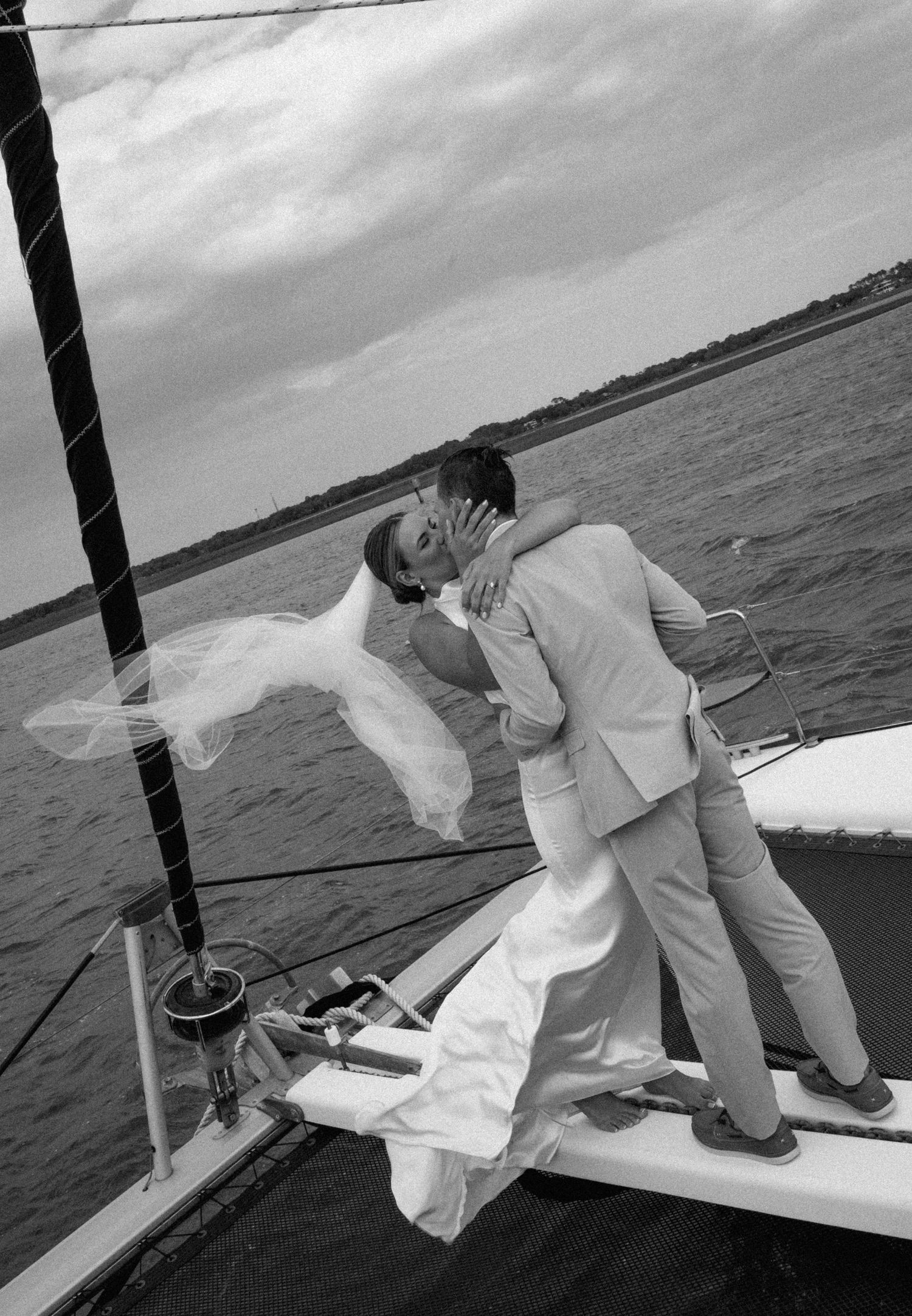 Black and white photo of bride and groom kissing on a sailboat with veil blowing in the wind.