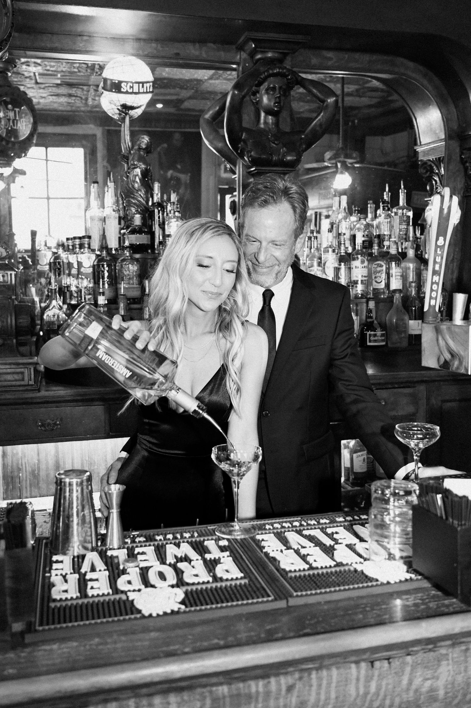Black and white photo of couple at a bar, woman pouring a drink while man embraces her.
