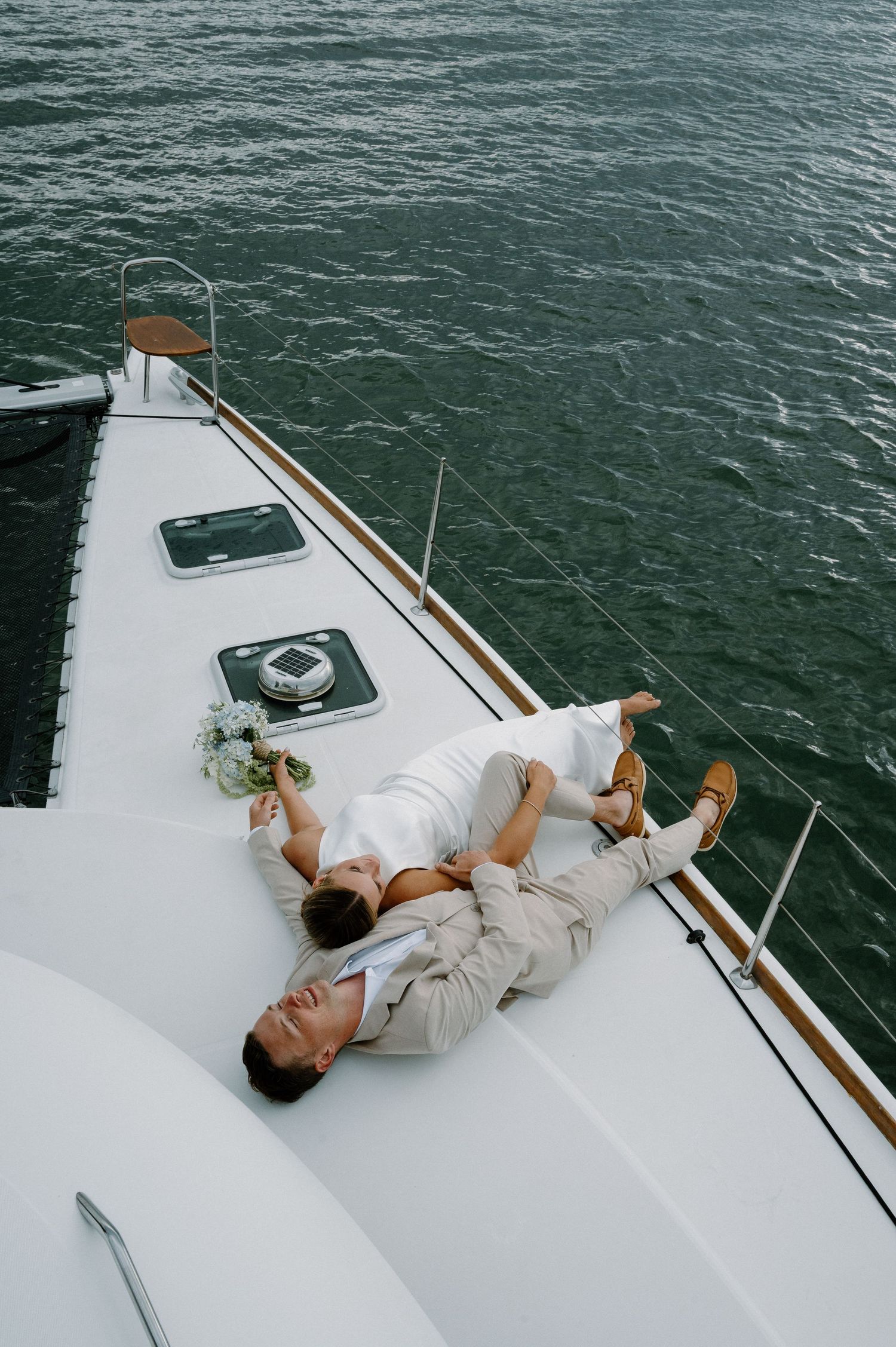 Bride and groom lying together on white catamaran deck with bouquet and ocean backdrop.